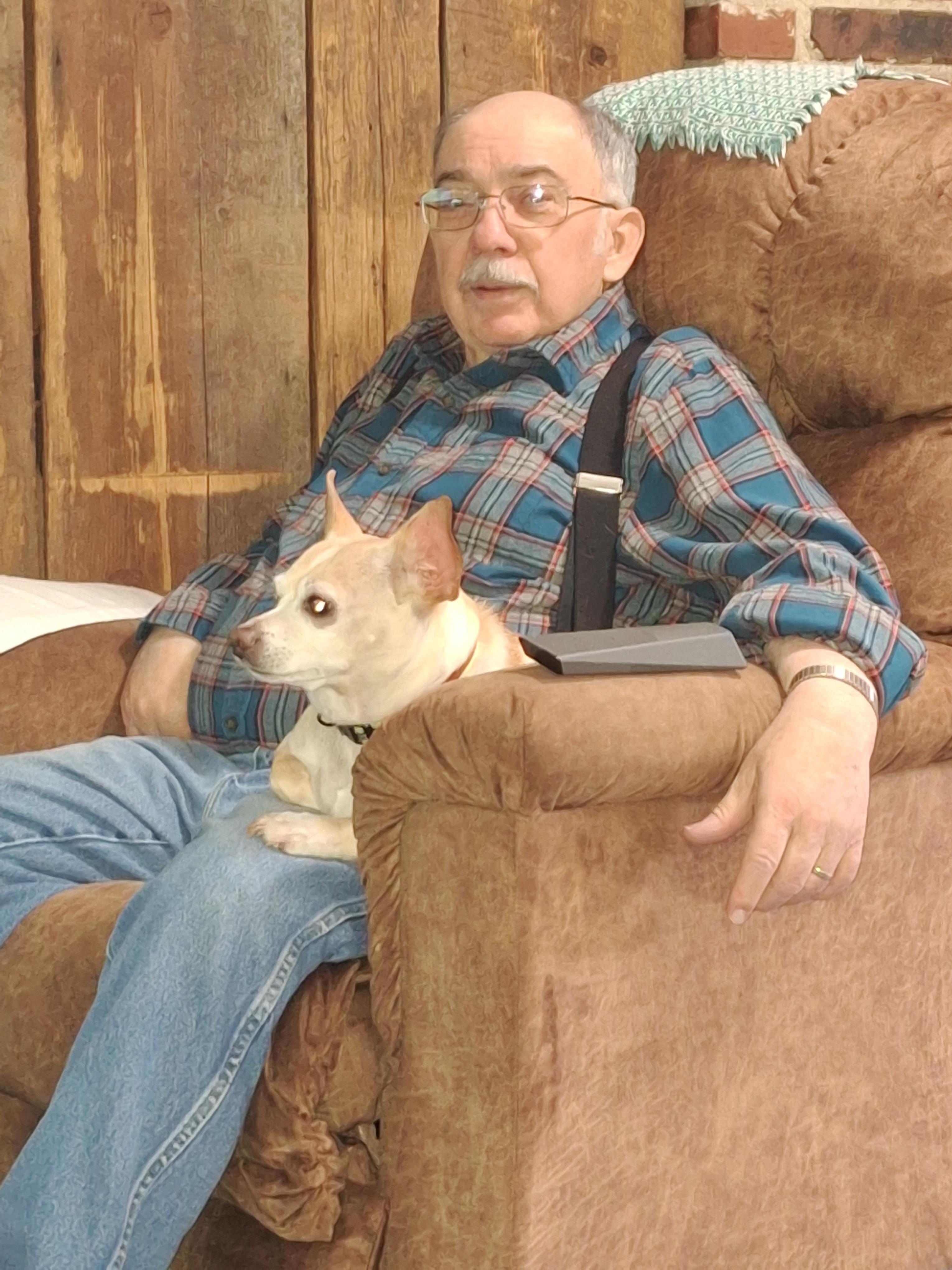 An elderly man sits comfortably in an armchair with his small dog beside him in a rustic space.
