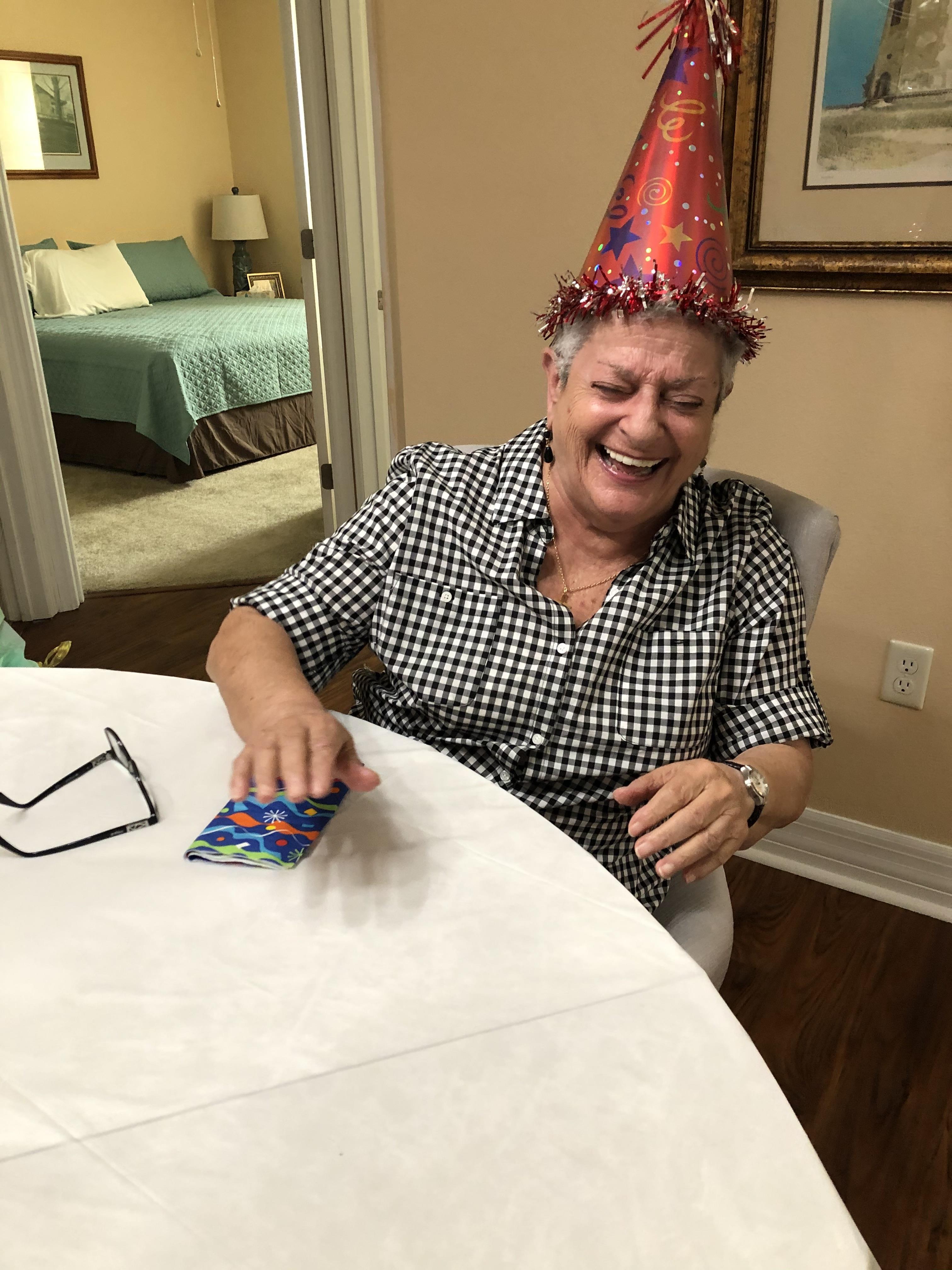 An elderly woman laughs heartily while wearing a festive party hat, surrounded by cheerful company.