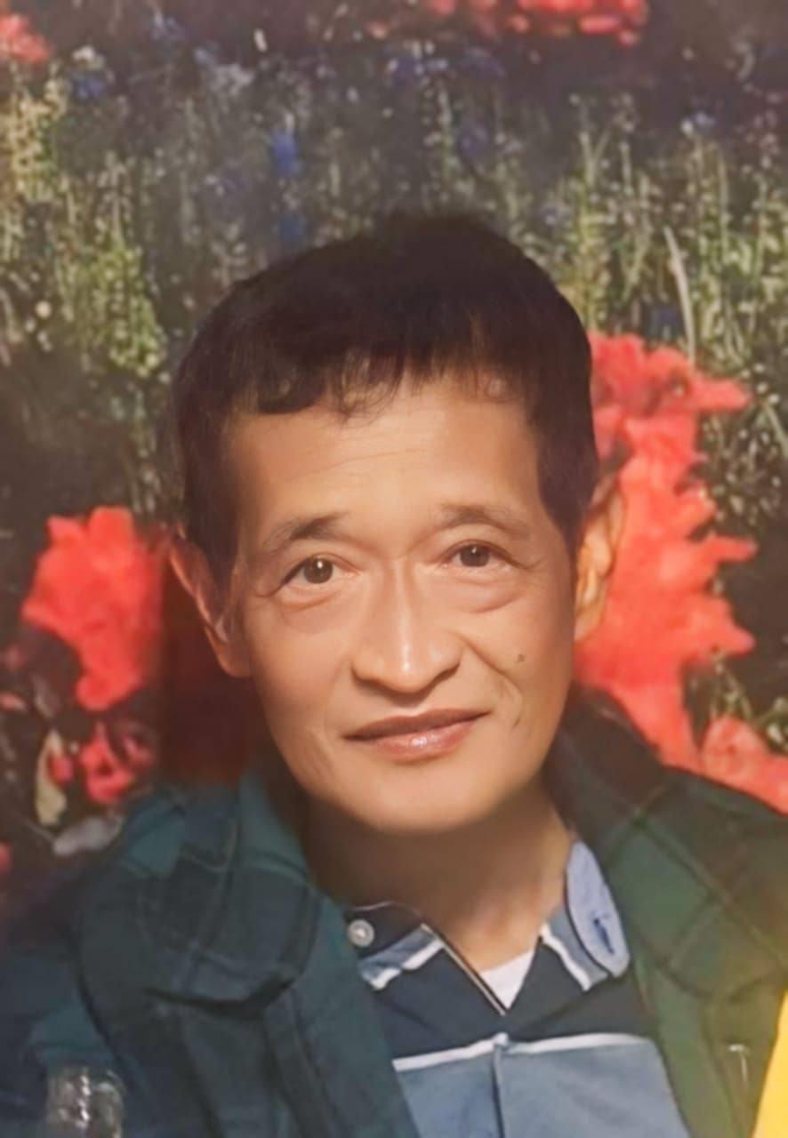 A smiling man poses in casual clothing with a bright floral backdrop, enjoying time with others.