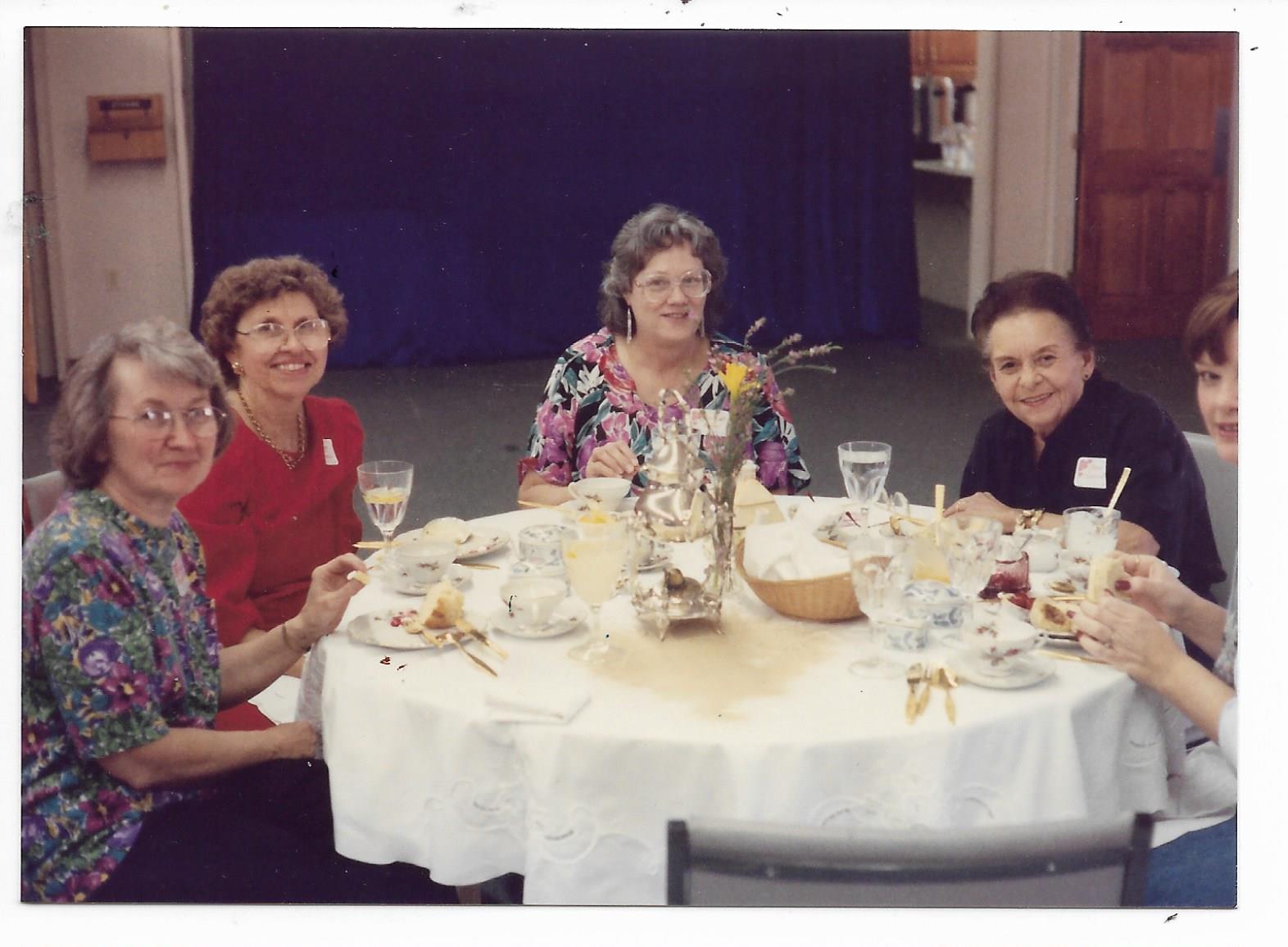 Five women share laughter and conversation while dining at a round table.