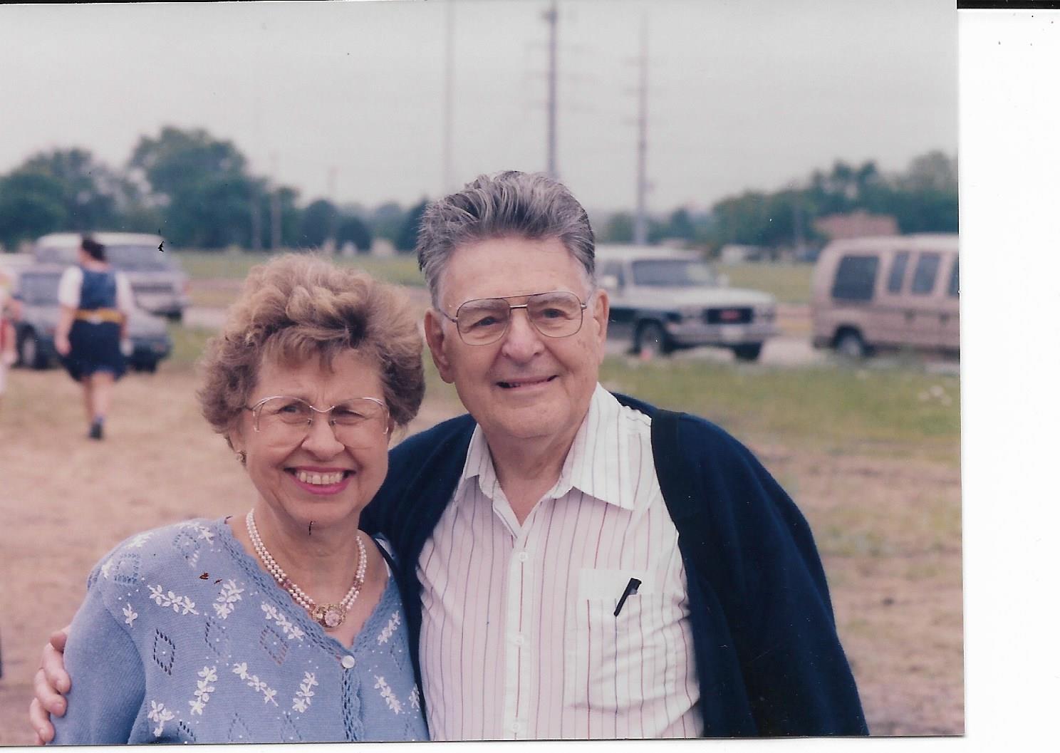 Couple smiles broadly together in a casual outdoor setting surrounded by vehicles and friends.