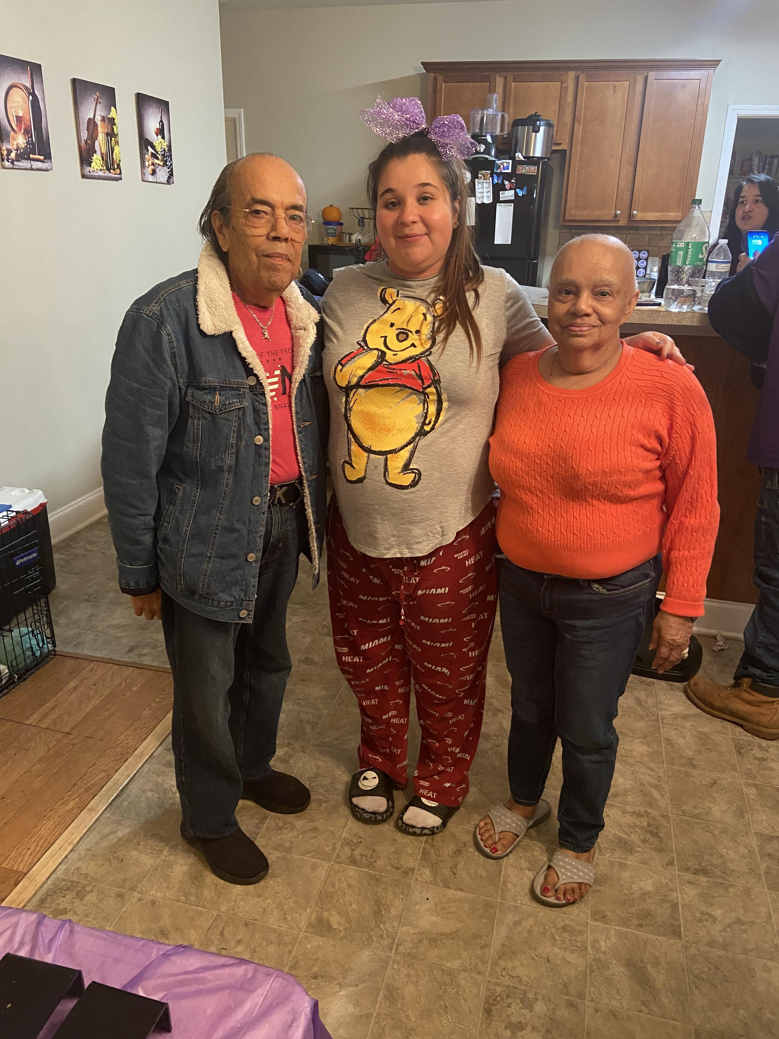 Three family members smile in a cozy living room, dressed casually for a gathering.