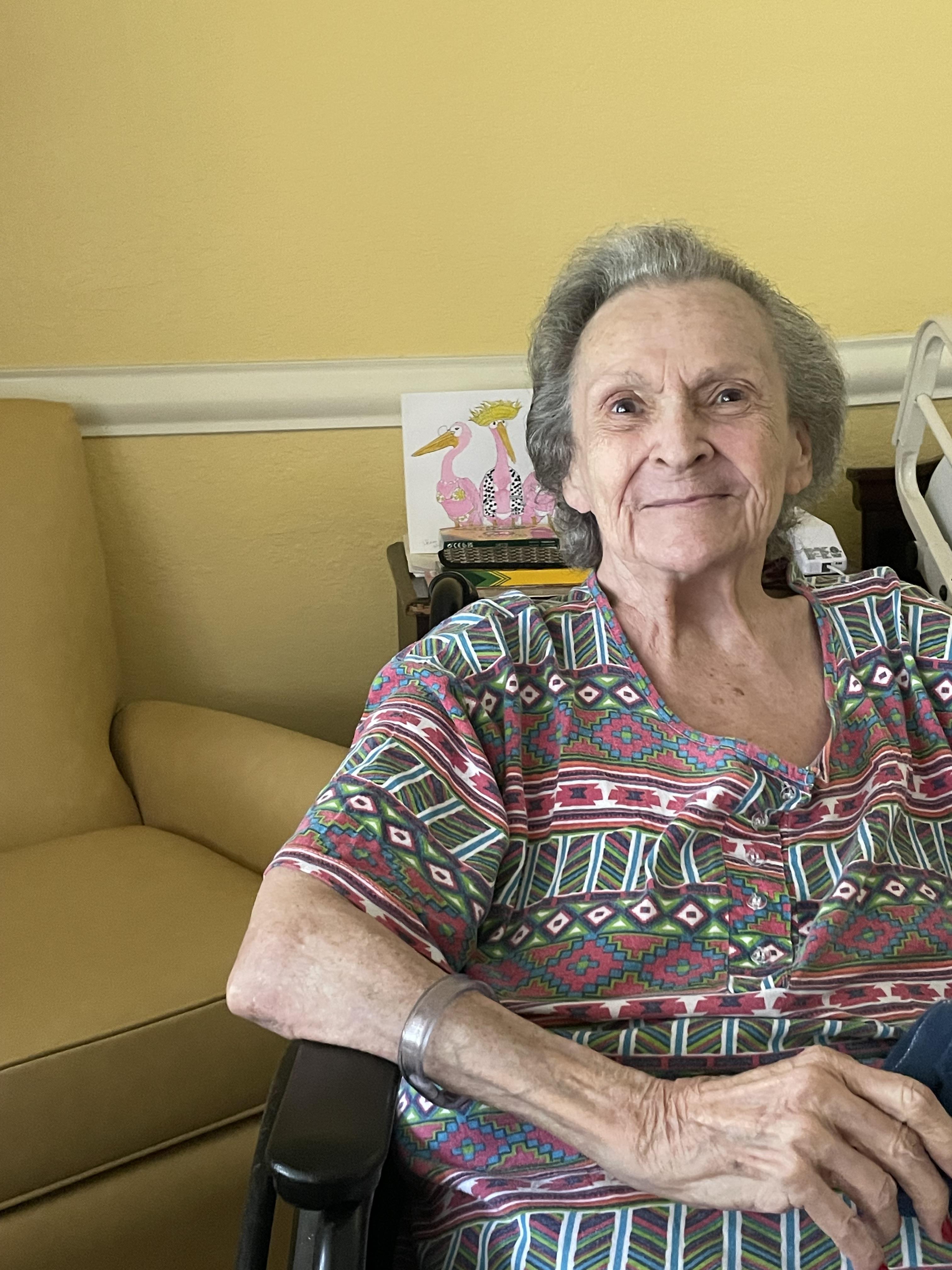 An elderly woman sits comfortably in a living room, smiling and enjoying her surroundings.