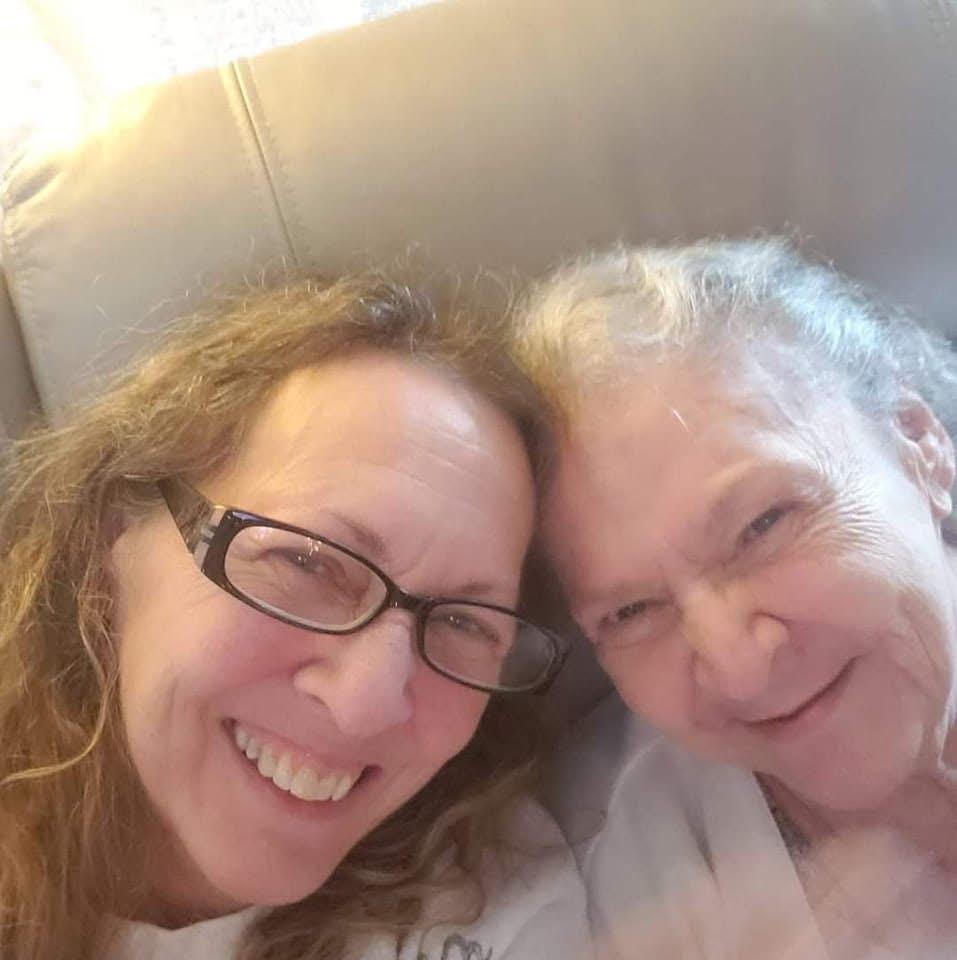 Two women smile widely, enjoying a close and happy moment while relaxing indoors.