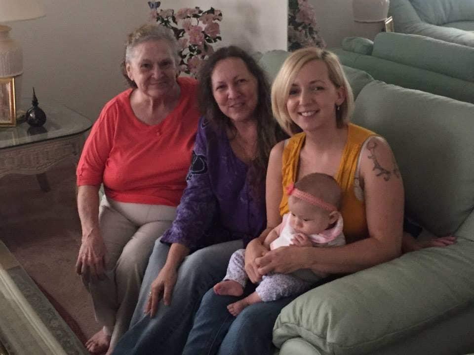 Three women, including a mother and grandmother, smile while holding a baby in a living room.