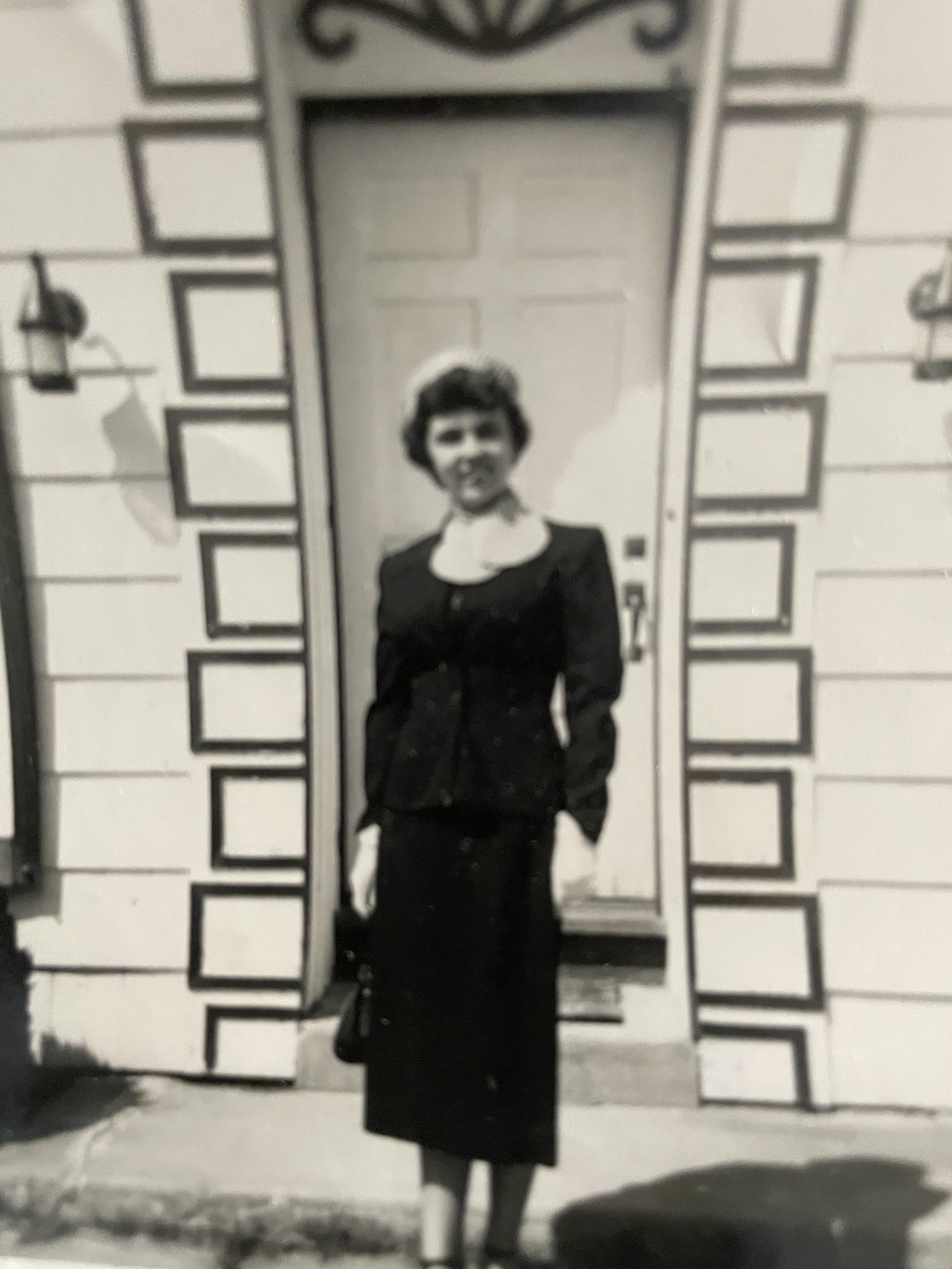 A woman smiles proudly in a stylish black outfit and pearl necklace outside a vintage building.