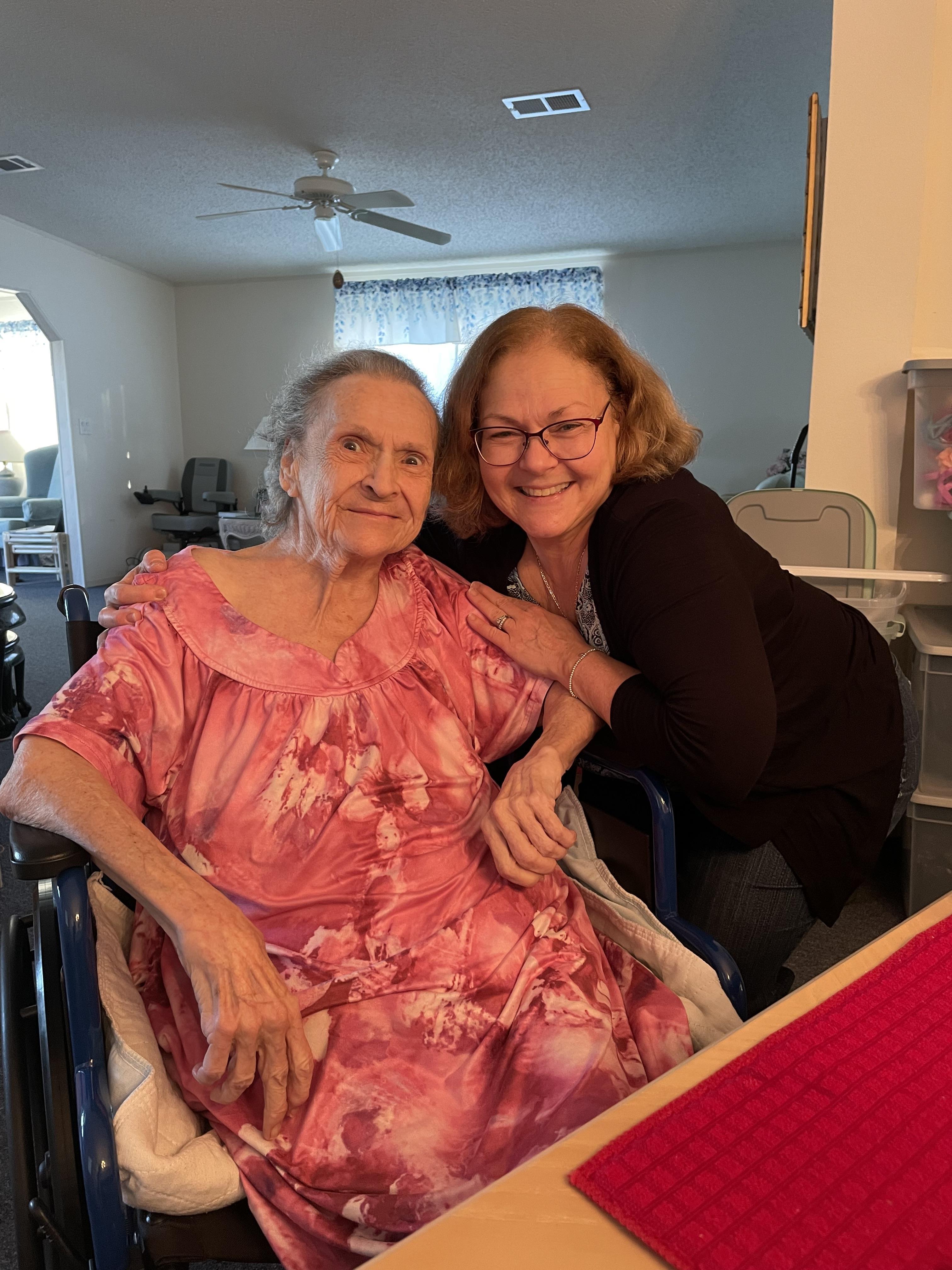 Senior resident wearing a floral dress enjoys time with a caregiver in a bright nursing home lounge.