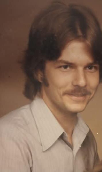 A young man with a mustache and long hair poses for a close-up portrait while smiling softly.