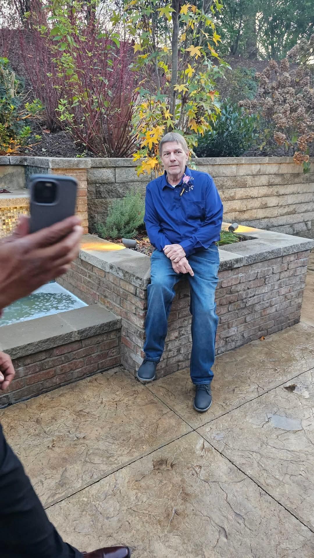 Individual casually seated on a stone bench with vibrant plants in the background.