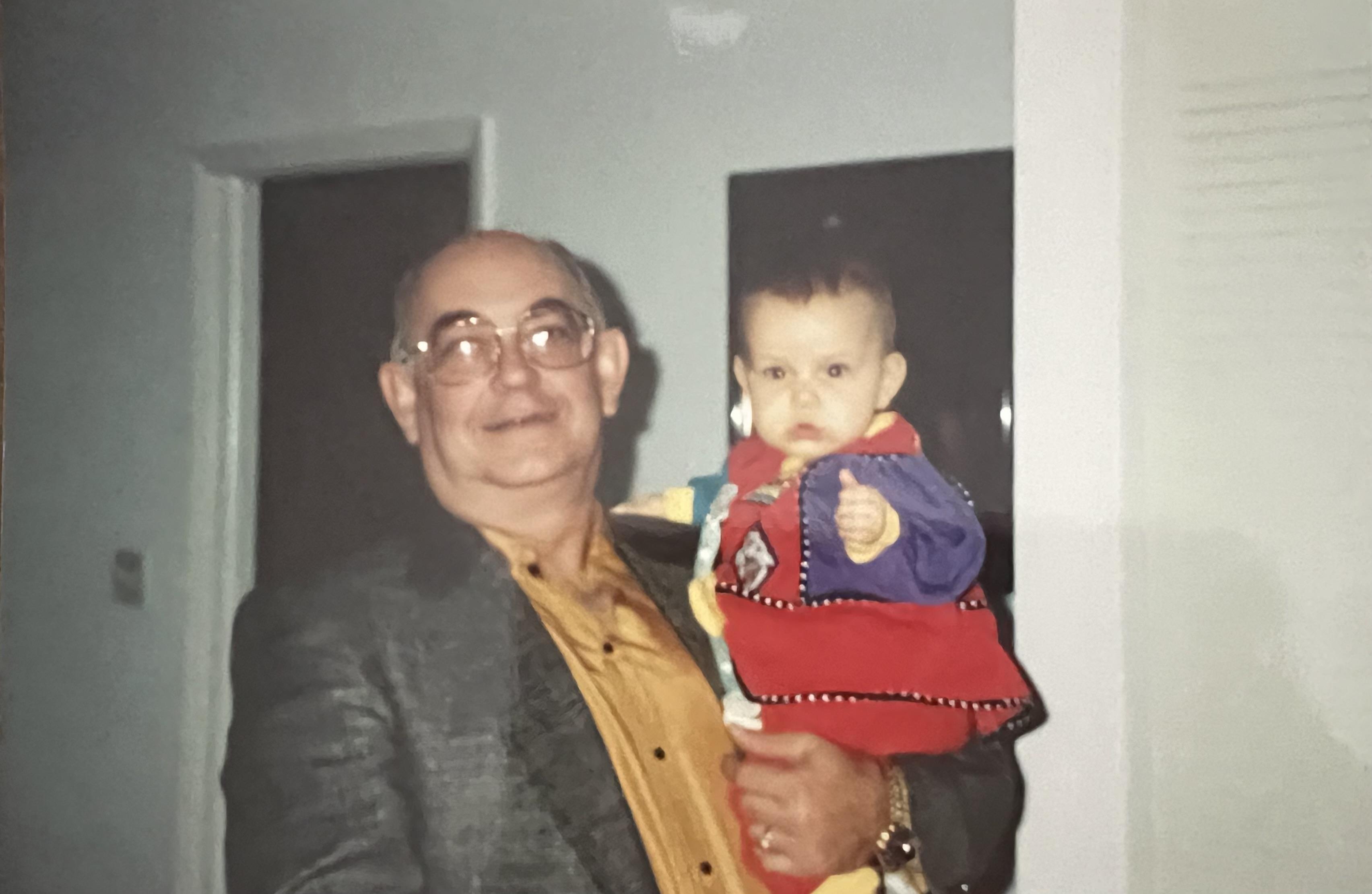 An elderly man smiles while holding a baby dressed in colorful clothing.