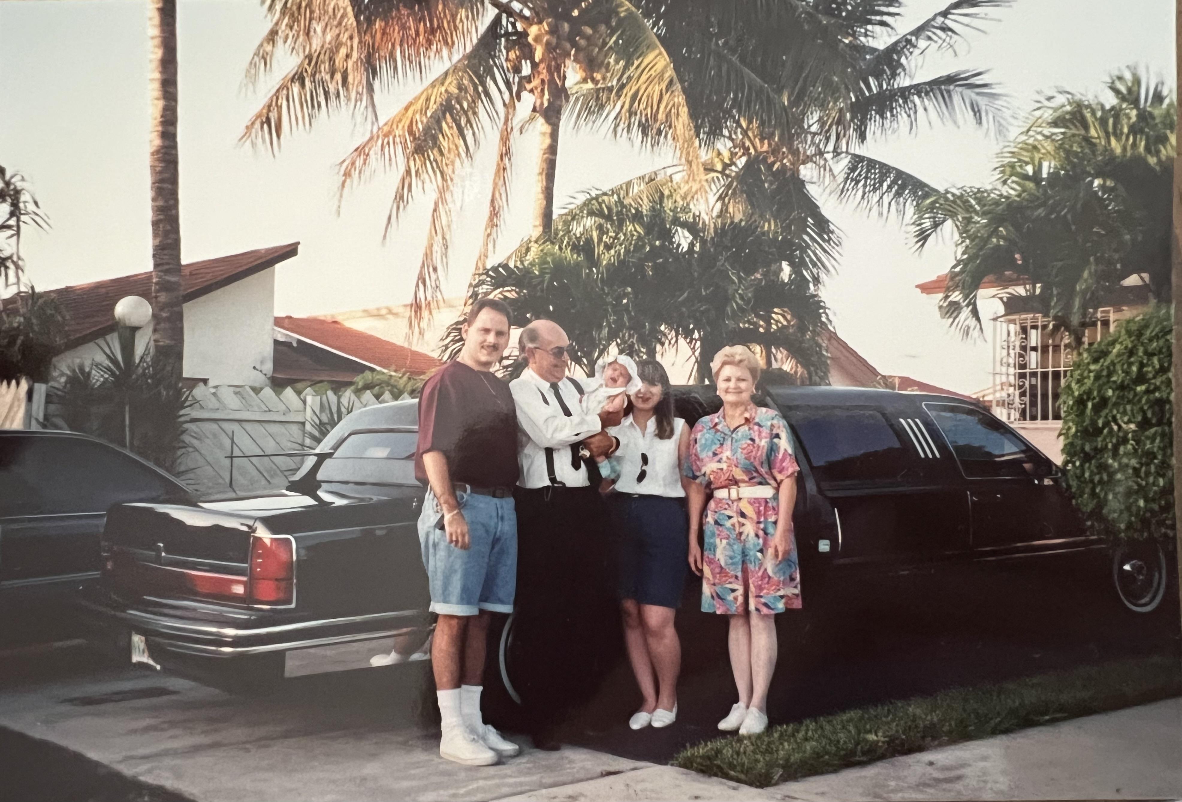 Four individuals are posing cheerfully beside a classic vehicle in a warm, tropical setting.