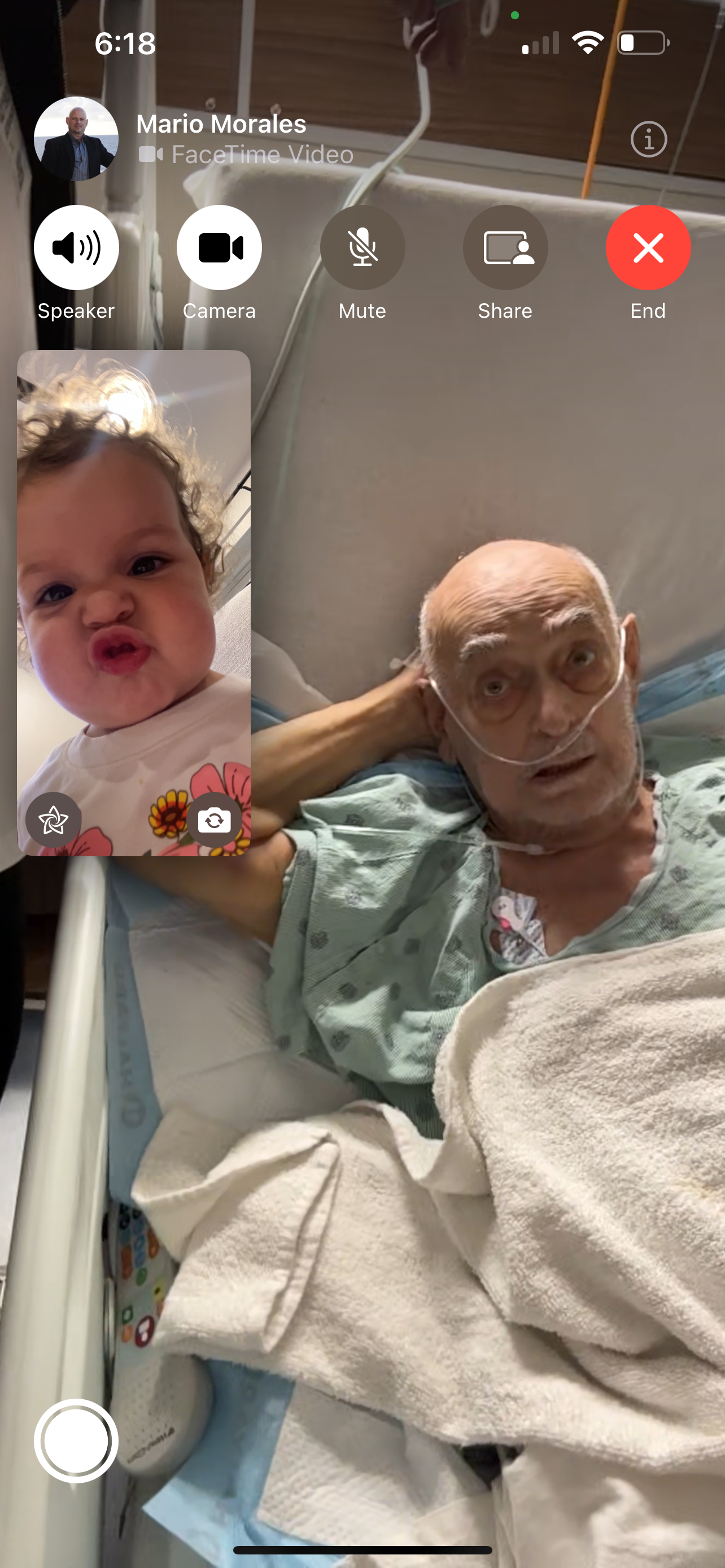 An elderly man rests in a hospital bed while video chatting with a baby on screen.