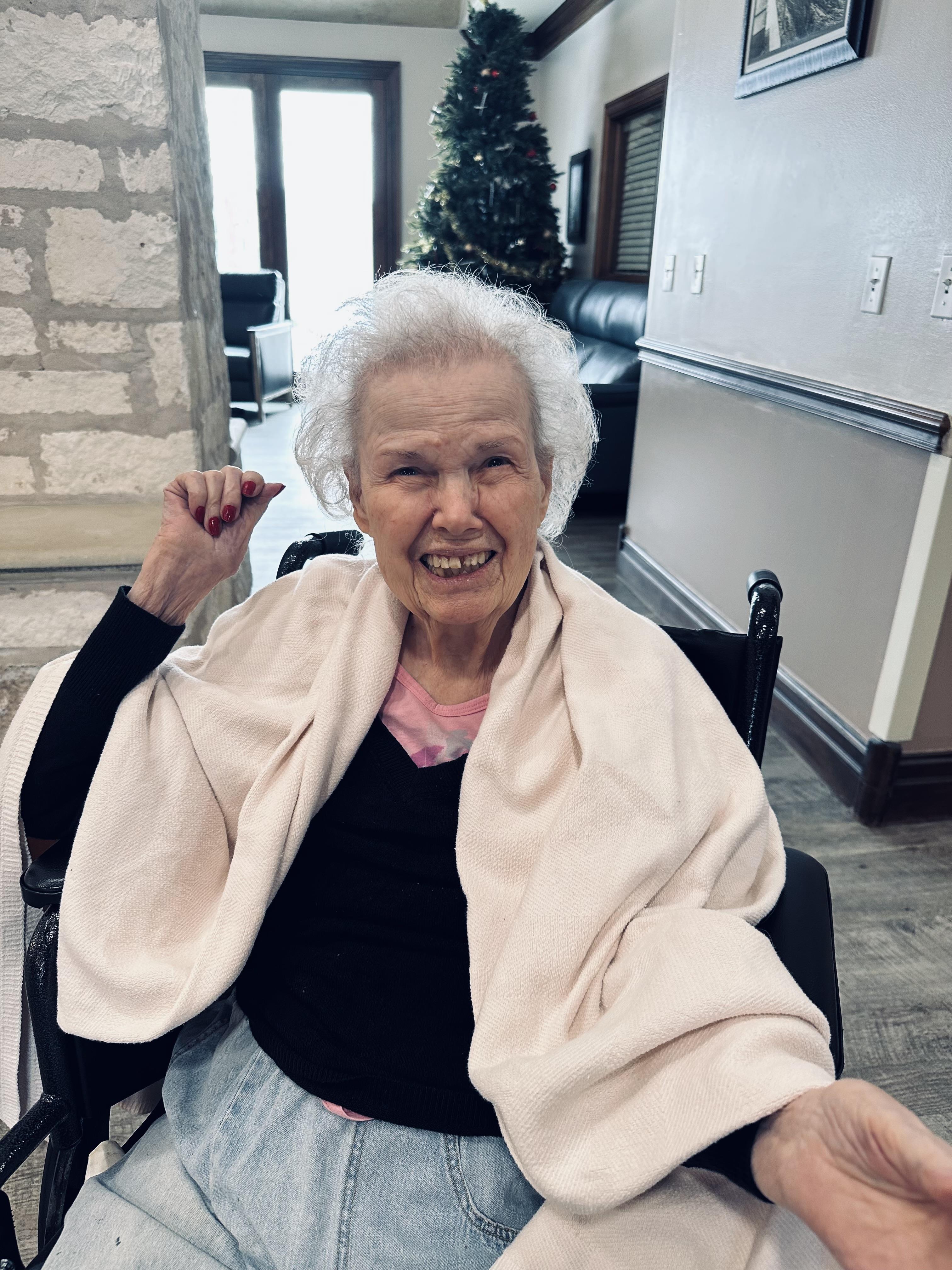 Elderly woman enjoys her time indoors, wrapped in a shawl, smiling cheerfully in a warm atmosphere.