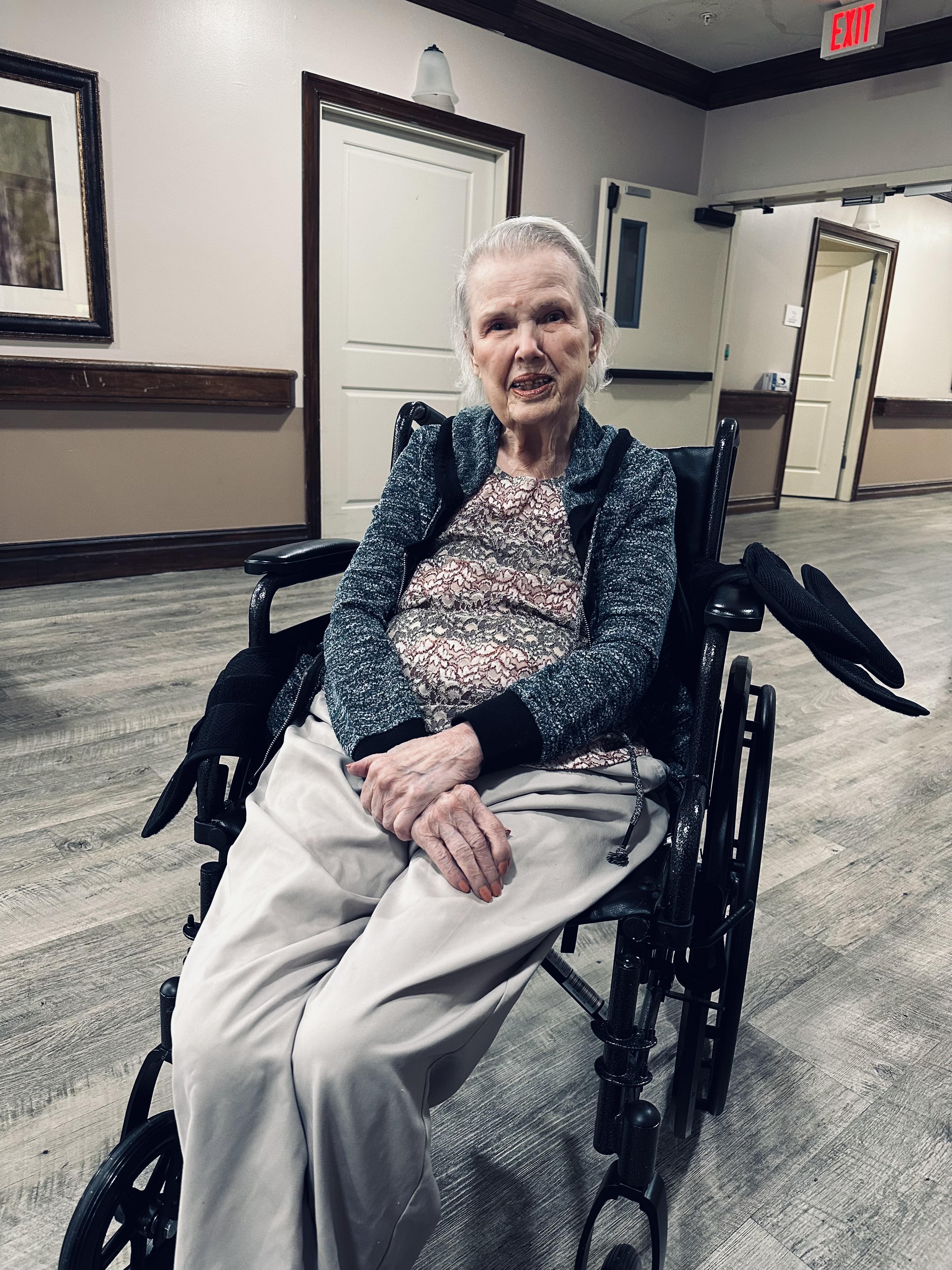 An elderly woman sits in a wheelchair, smiling peacefully in a nursing home hallway.