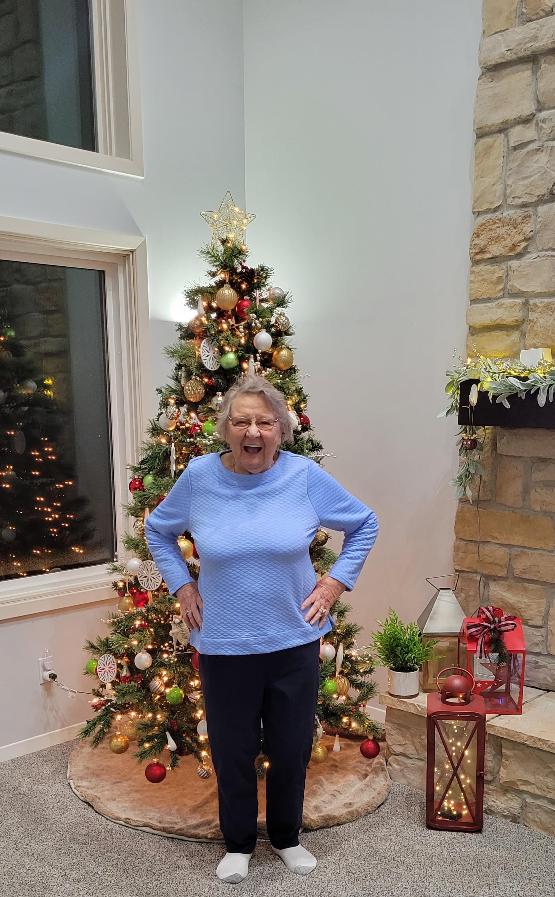 An elderly woman smiles joyfully in front of a beautifully decorated Christmas tree.
