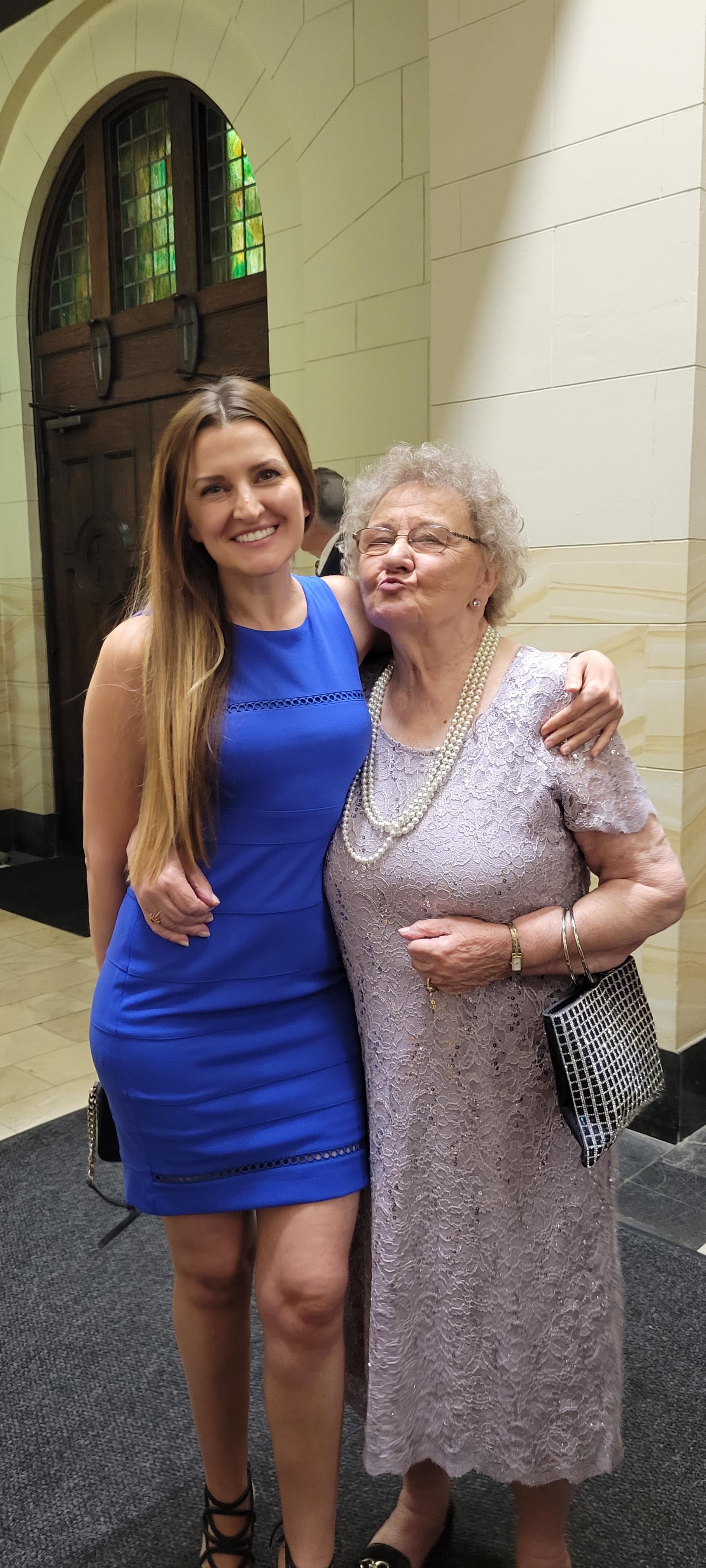 A young woman in a blue dress stands beside an elderly lady in a lavender outfit, smiling.