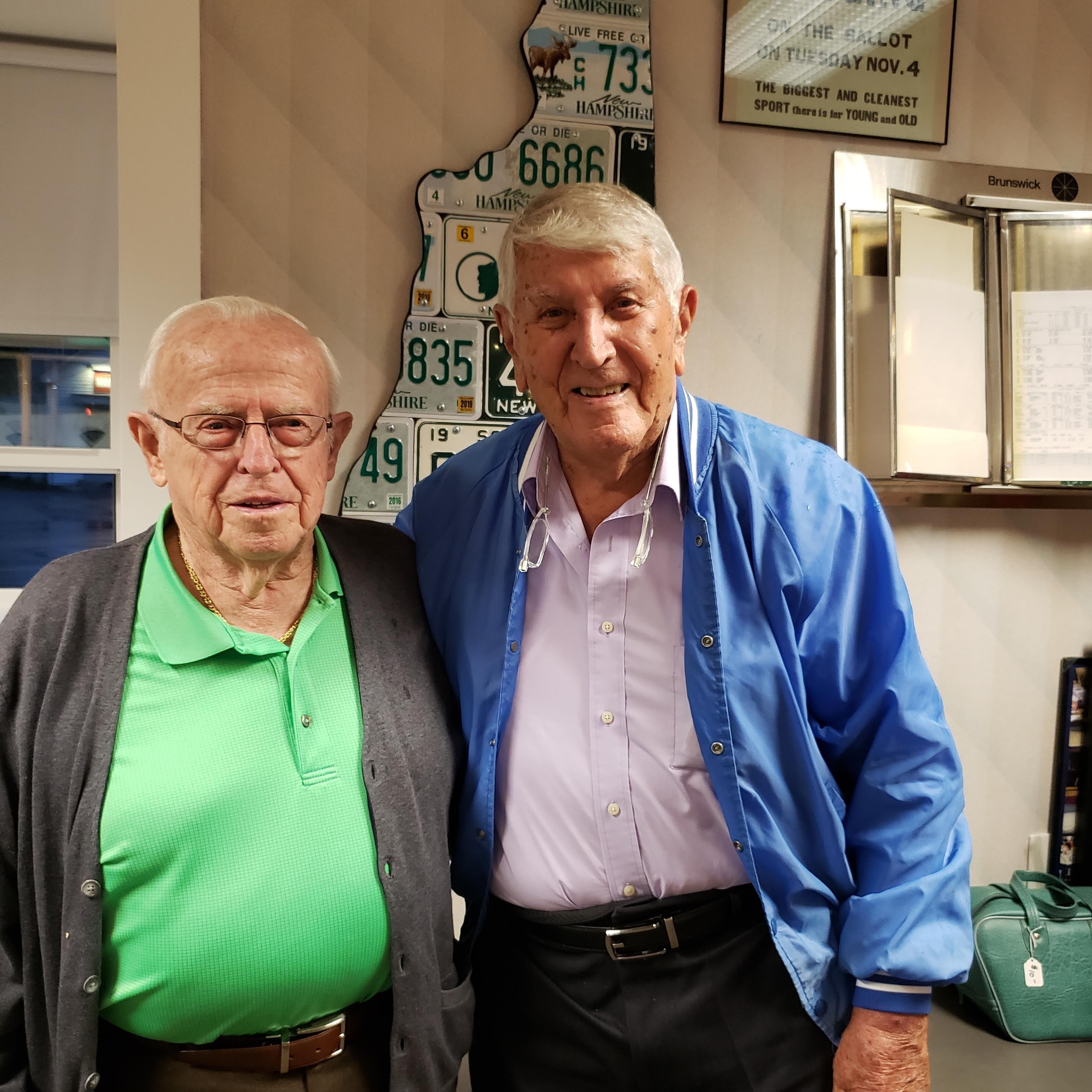 Two elderly men smile together in a cozy room filled with memorabilia.