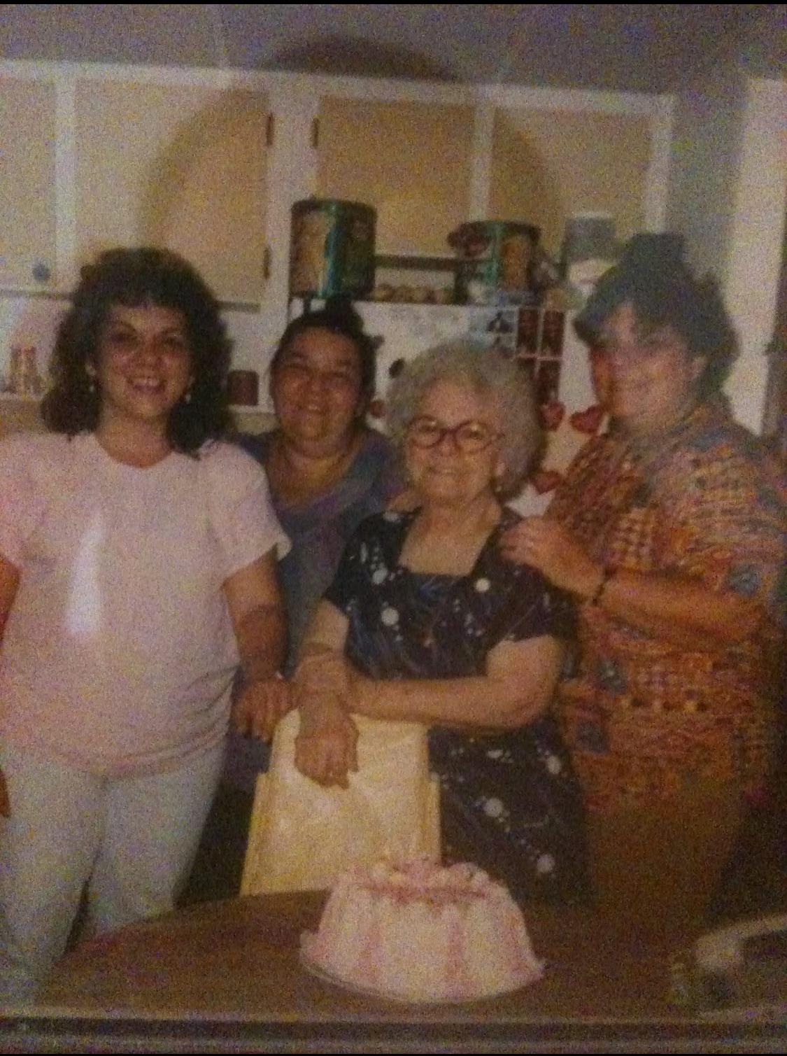 Four generations of family members enjoy a celebration in the kitchen with a cake.