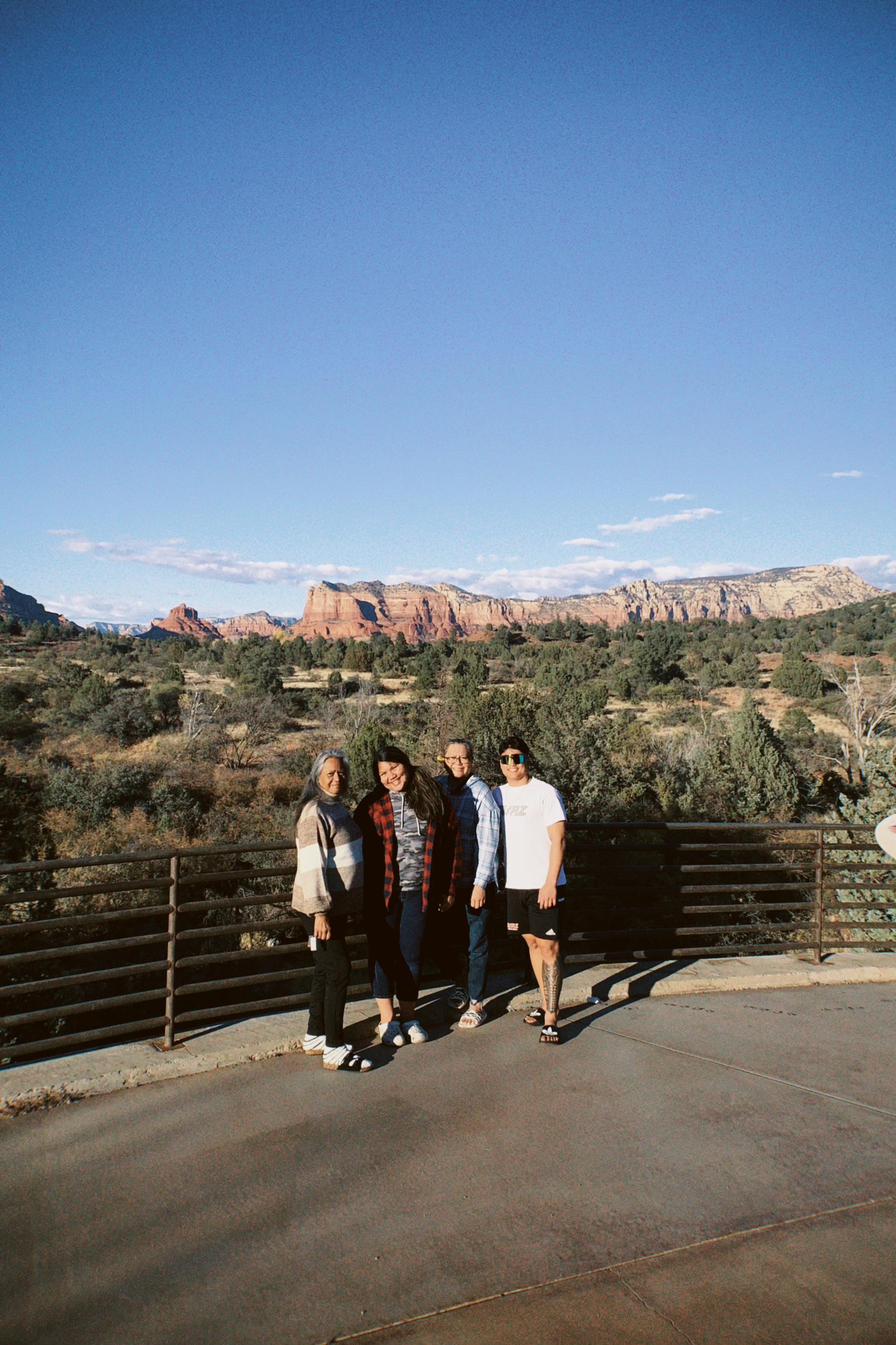 Group of friends stands in front of beautiful red rock formations under a clear blue sky.