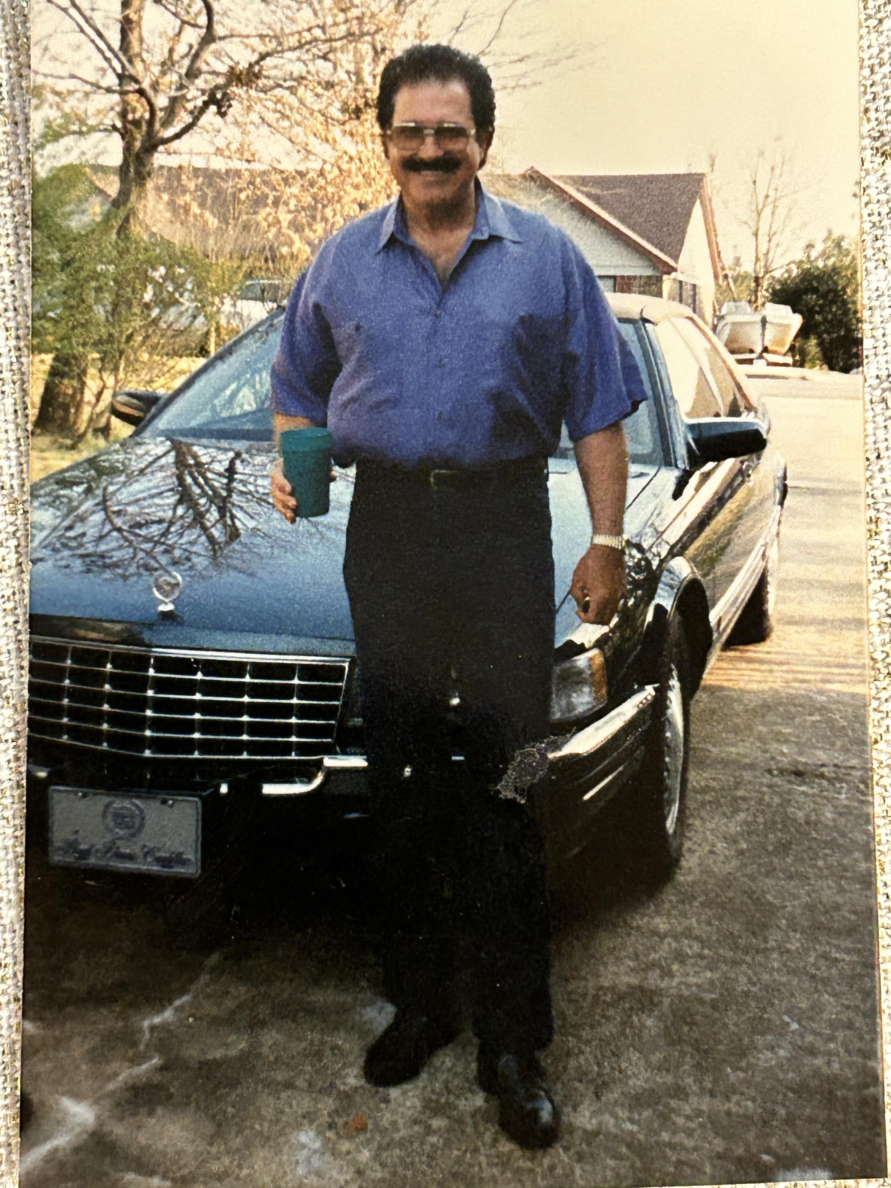 A man with a mustache wears a blue shirt and gray pants next to a black luxury vehicle.