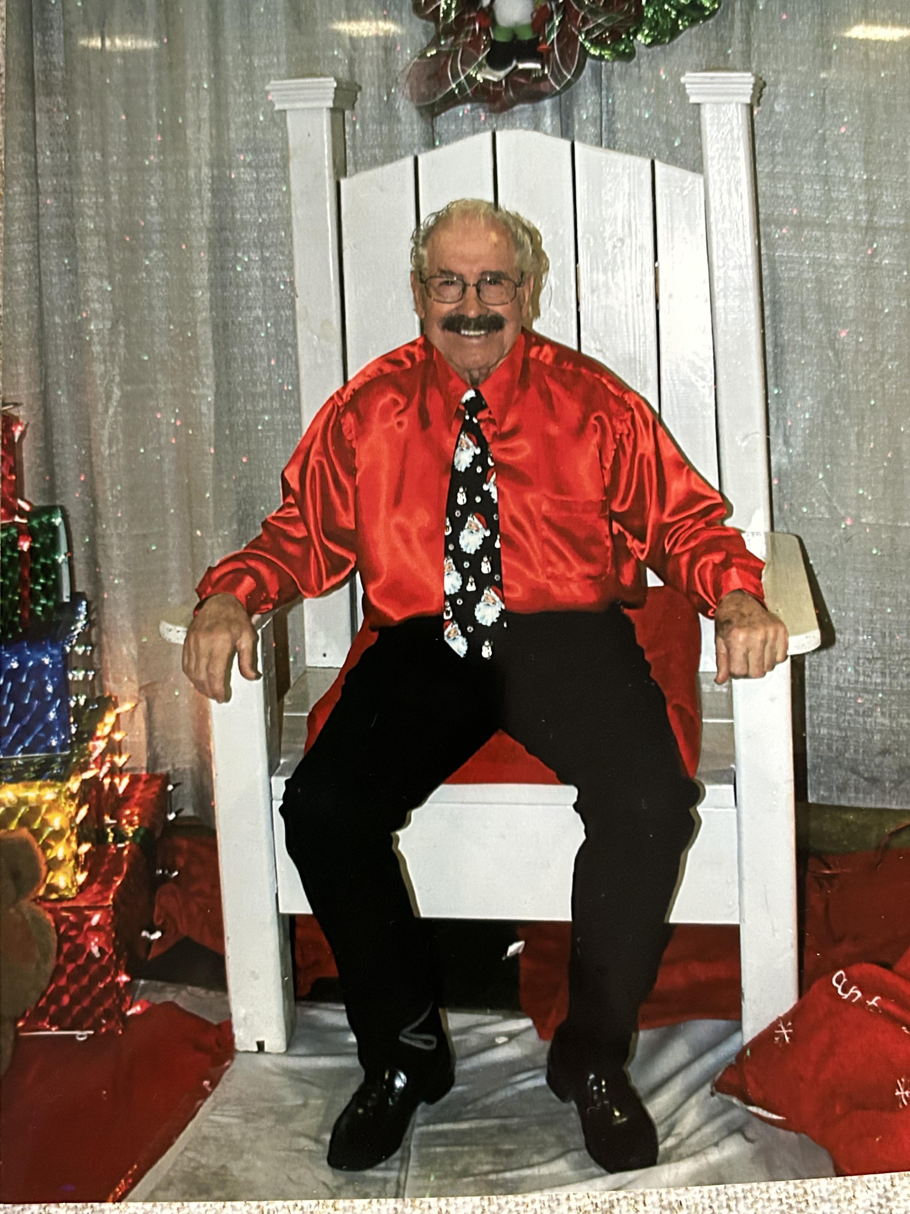 An elderly man in a red shirt and festive tie sits in a large chair at a holiday event.