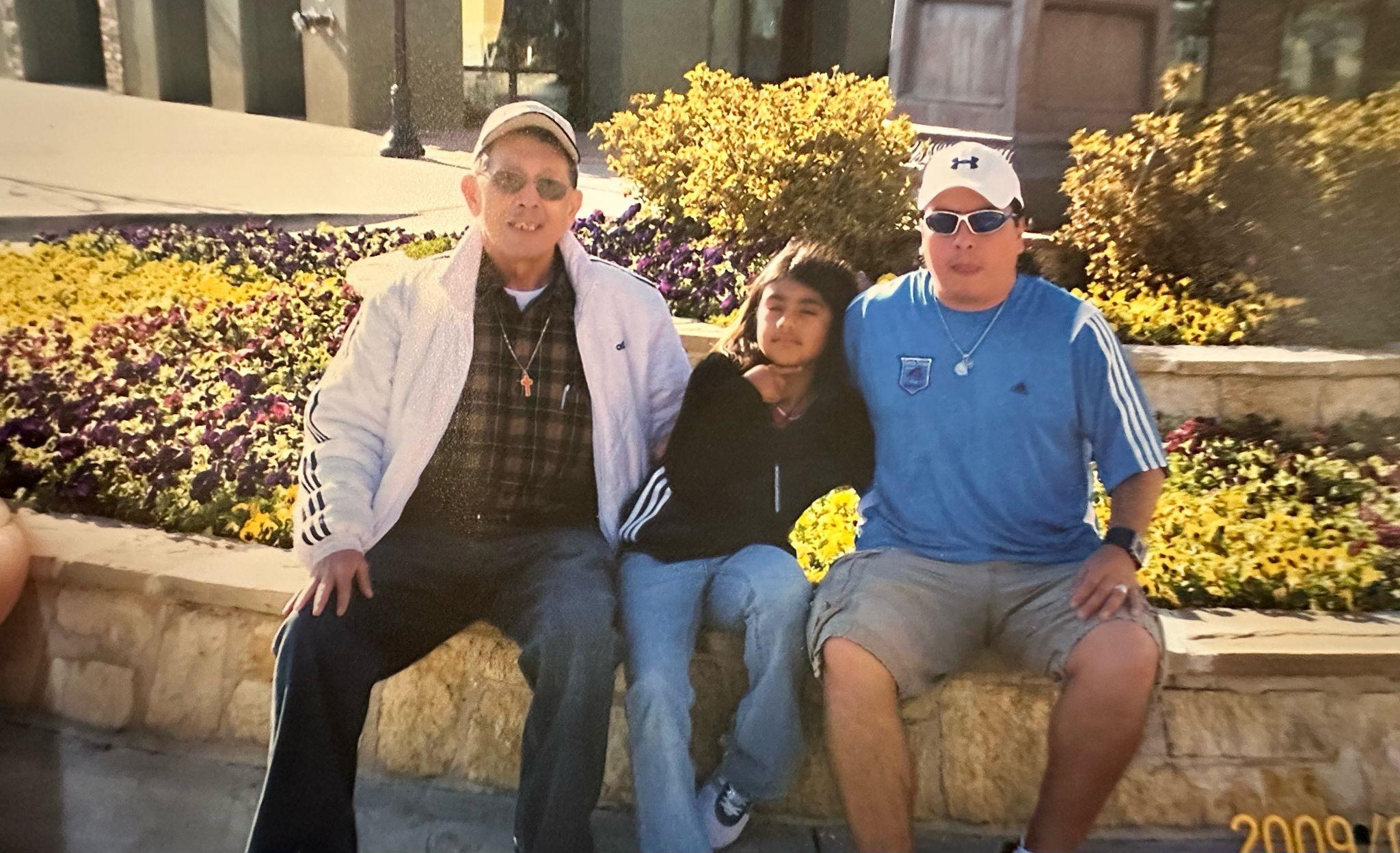 Three friends sit closely on a stone bench surrounded by colorful flowers in a park.