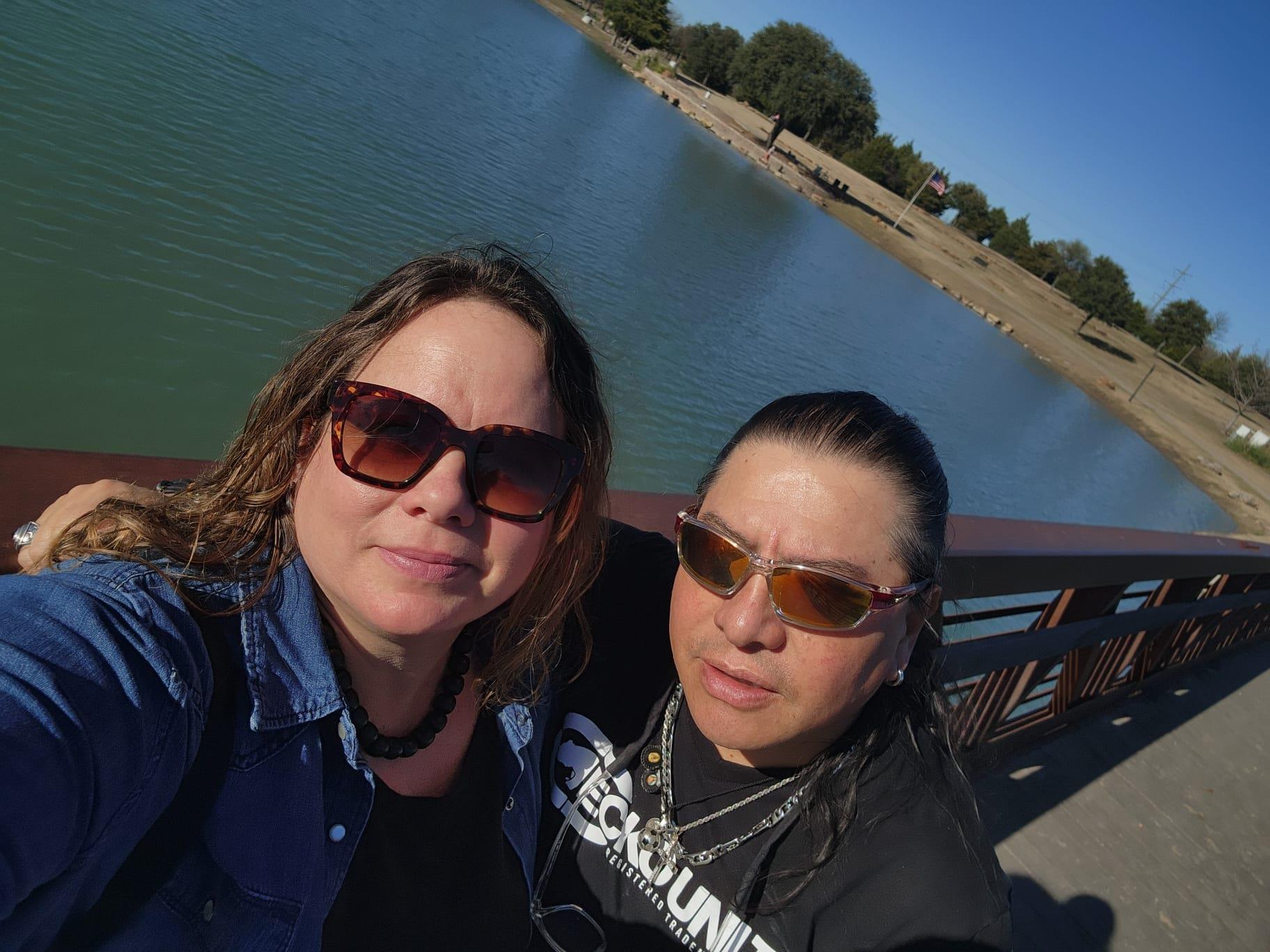 Two friends smile and pose together near a serene lake on a clear day.