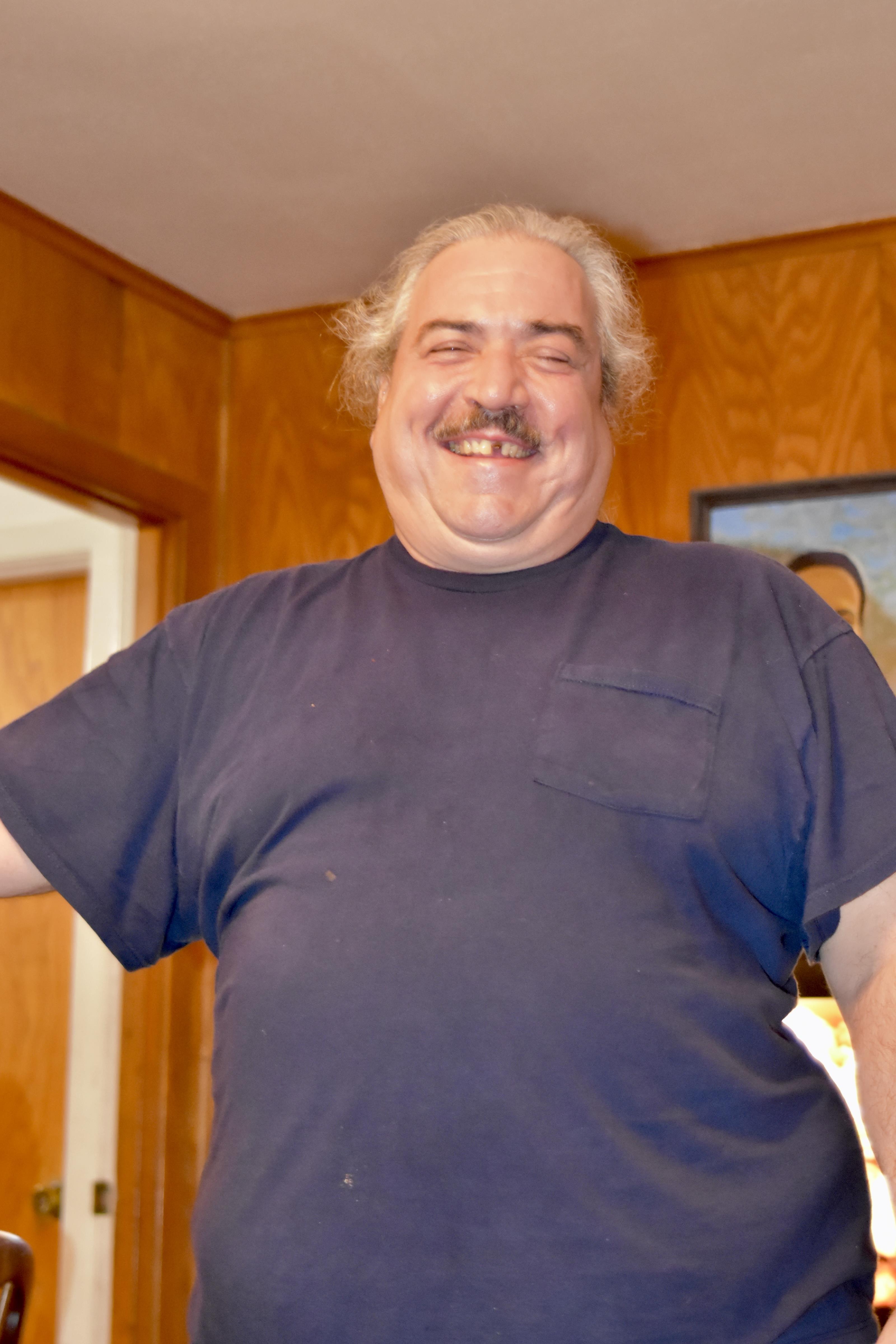 A cheerful man stands with a big smile in a warm, inviting room decorated with wooden accents.