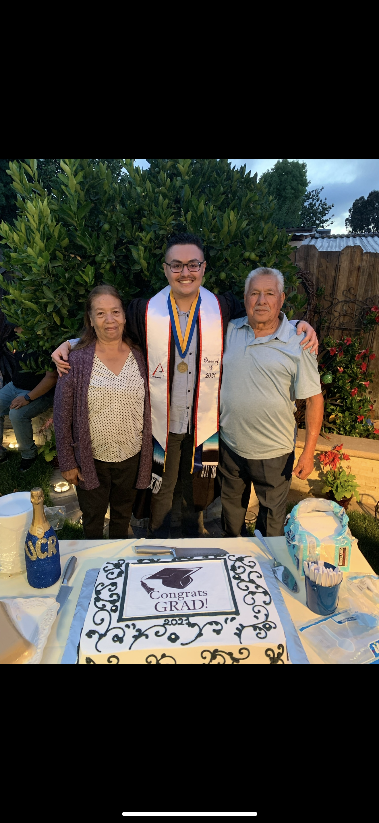 A graduate poses with grandparents by a cake during a festive outdoor celebration at dusk.
