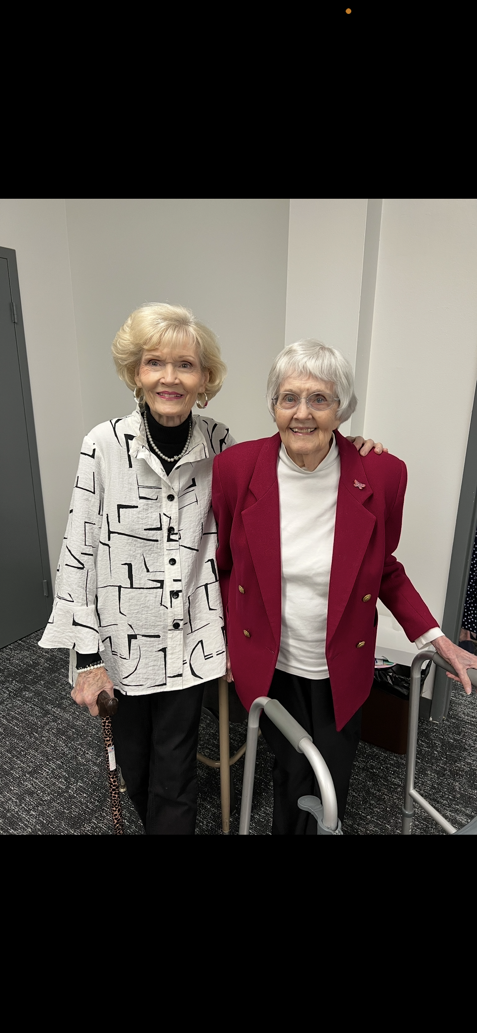 Two elderly women stand side by side, smiling in stylish outfits, enjoying their time.