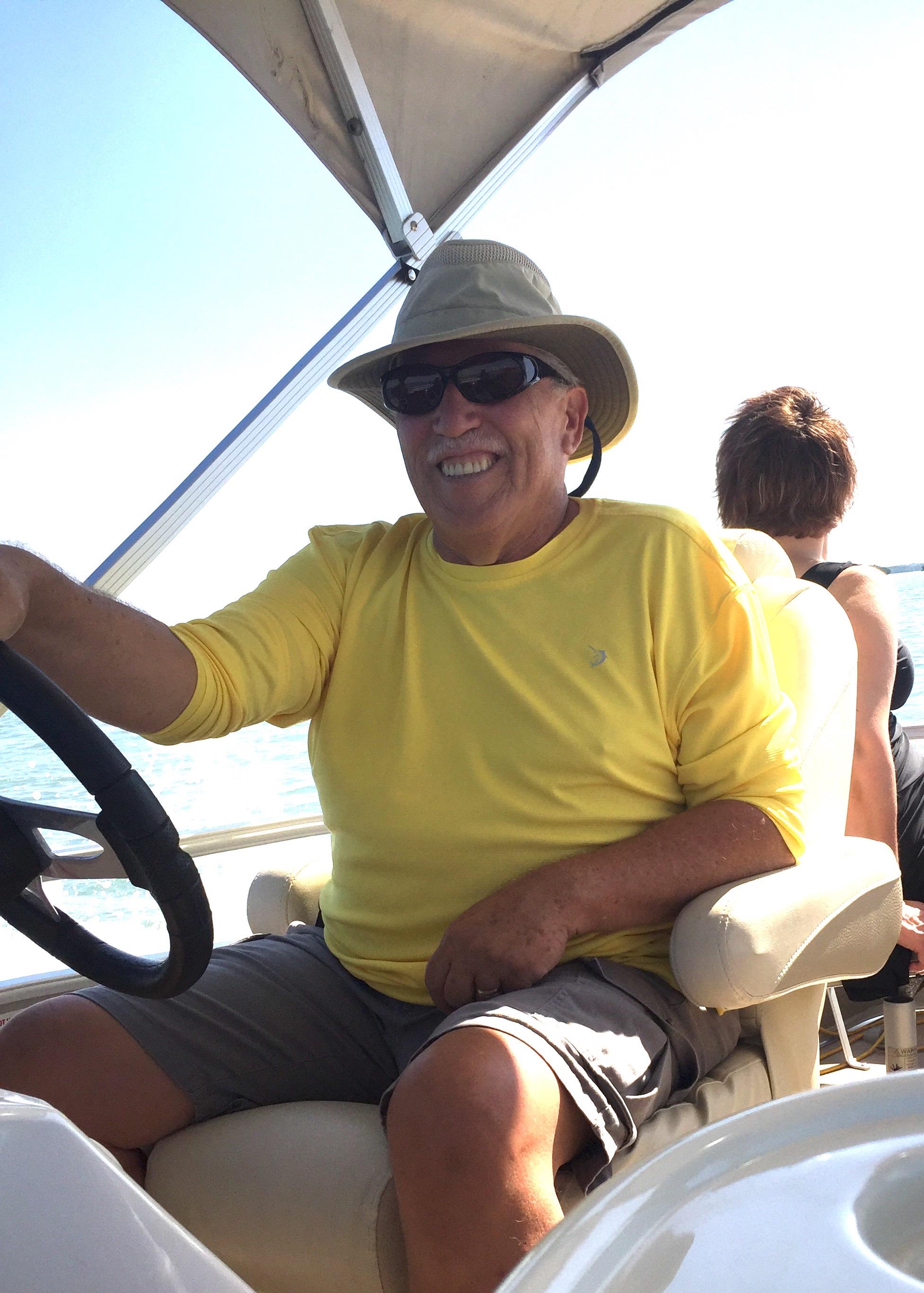 A man in a yellow shirt enjoys steering a boat with a smile while a woman relaxes behind him.