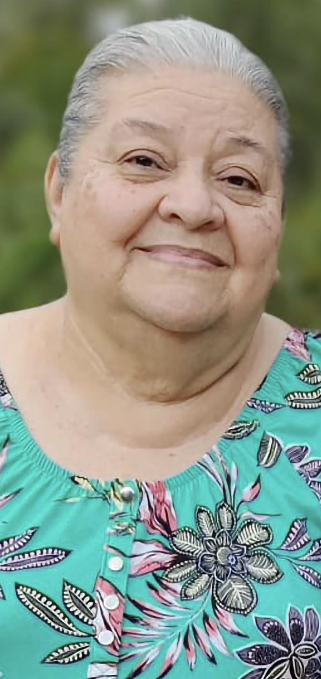 Elderly woman with a cheerful smile stands outside, wearing a colorful floral top on a bright day.