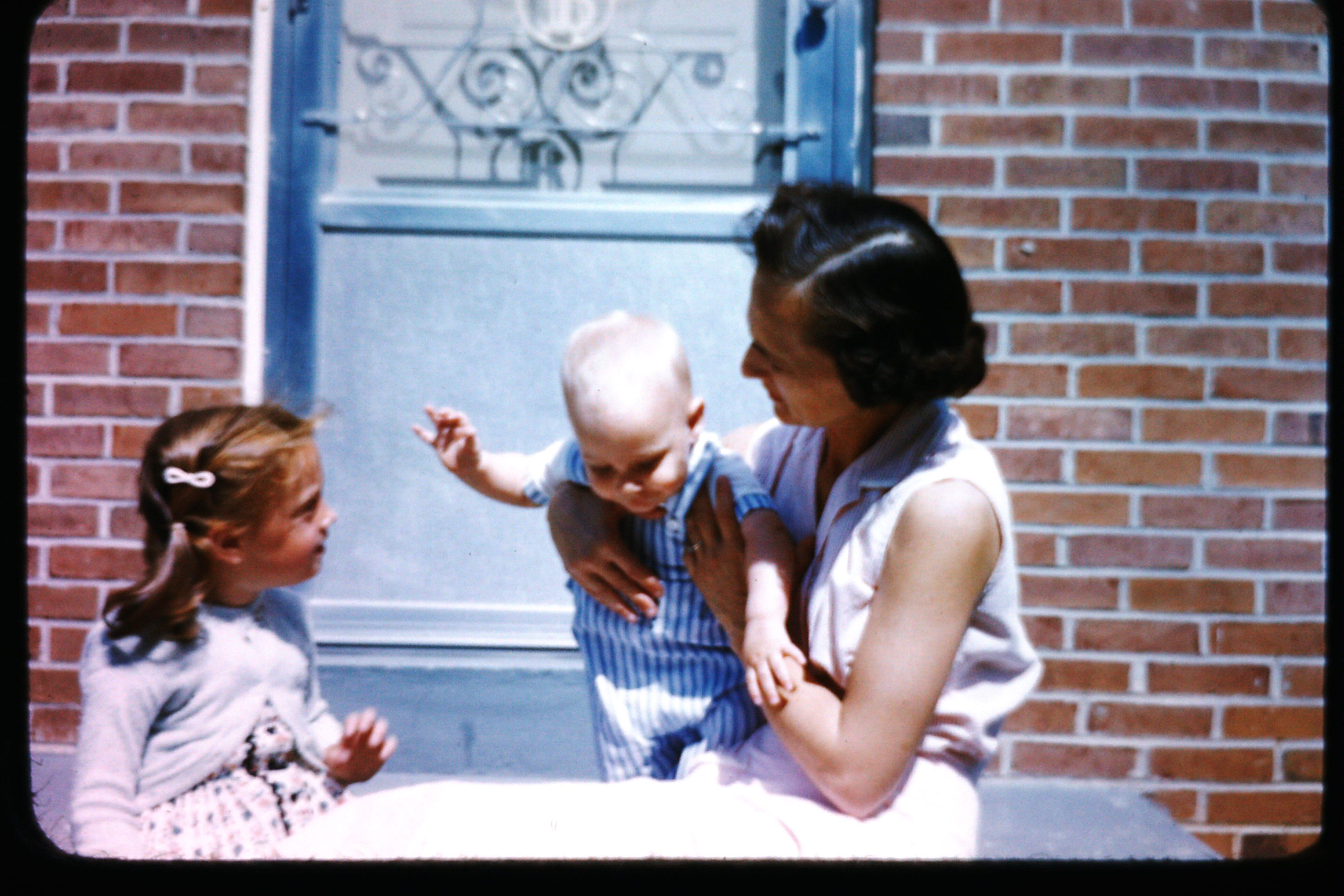 A mother holds her baby while a young girl engages in playful conversation nearby.