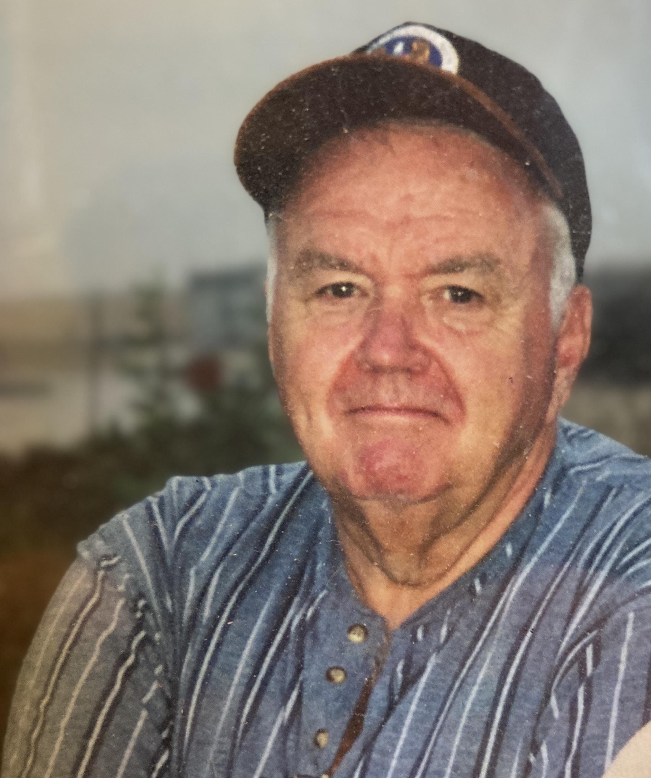 A senior man with a friendly demeanor poses outdoors wearing a baseball cap and casual shirt.