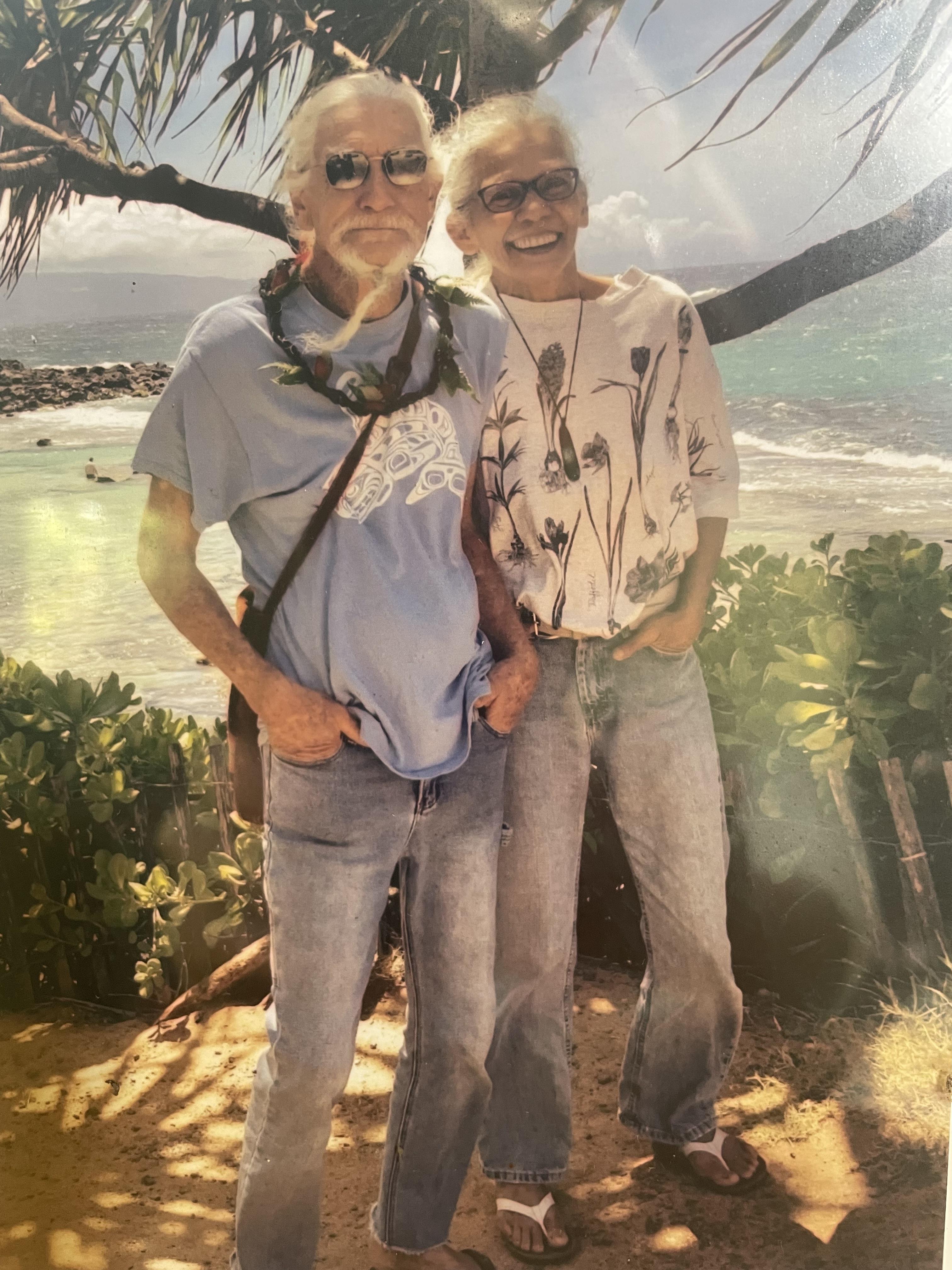 An elderly couple smiles brightly while standing near the shoreline under palm trees.