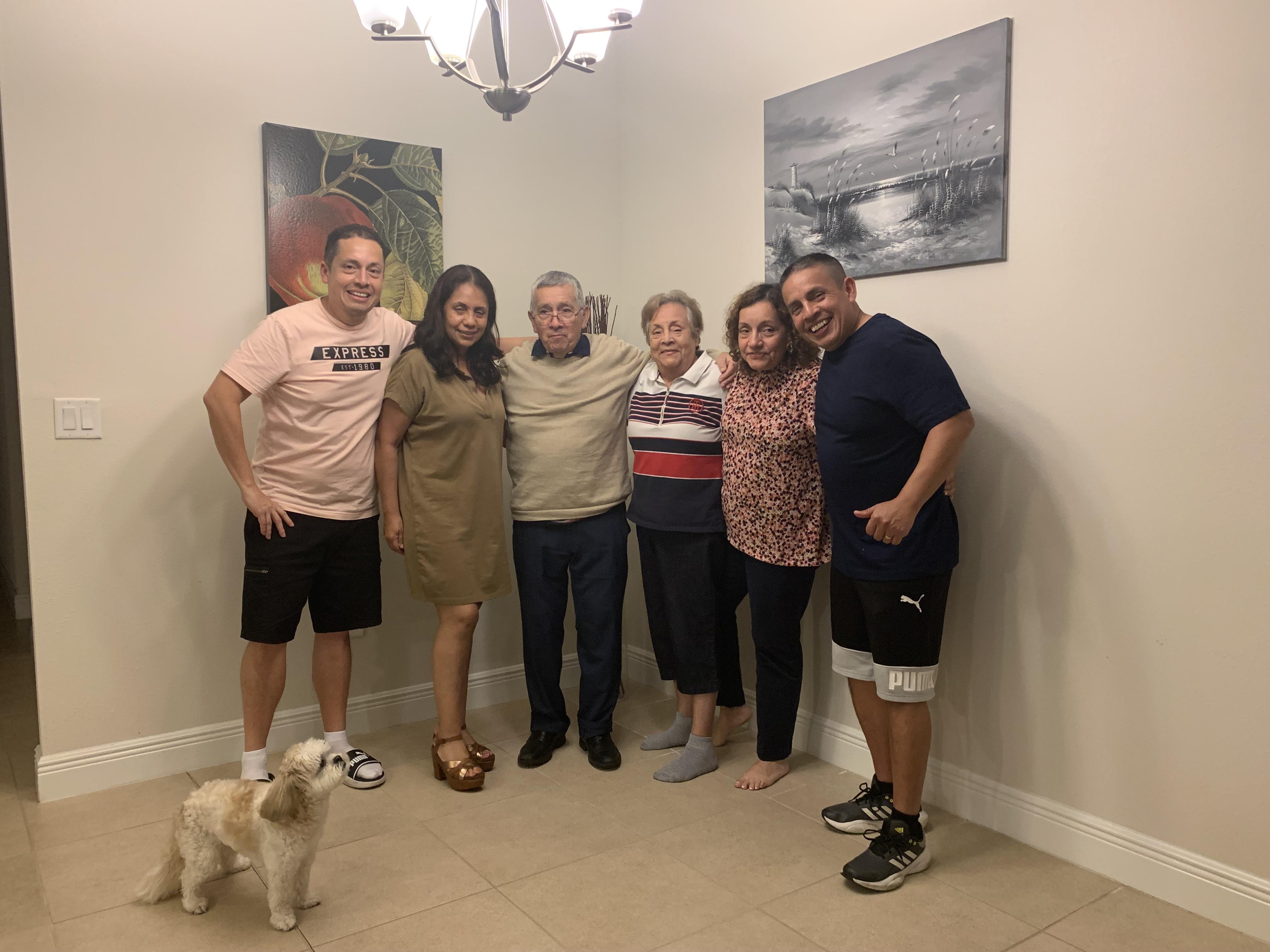 A group of six people stands together in a warm, inviting living room with a small dog.