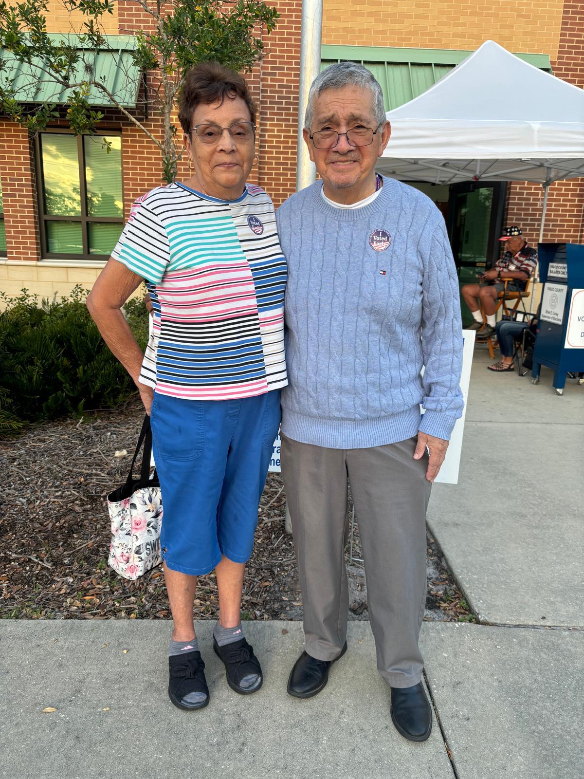 An elderly couple stands side by side outside a community event, smiling brightly.