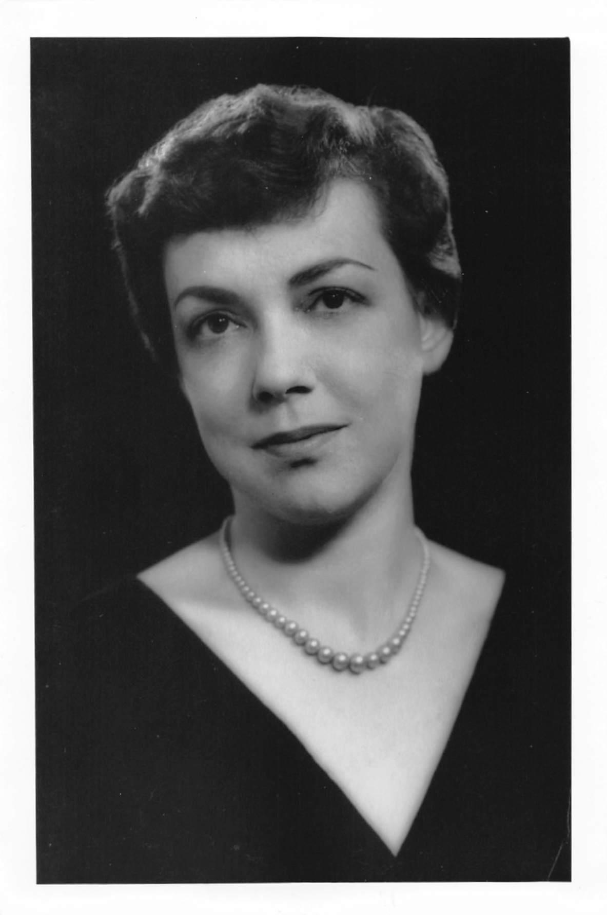 A woman poses elegantly with a confident expression, wearing a pearl necklace and dark attire.