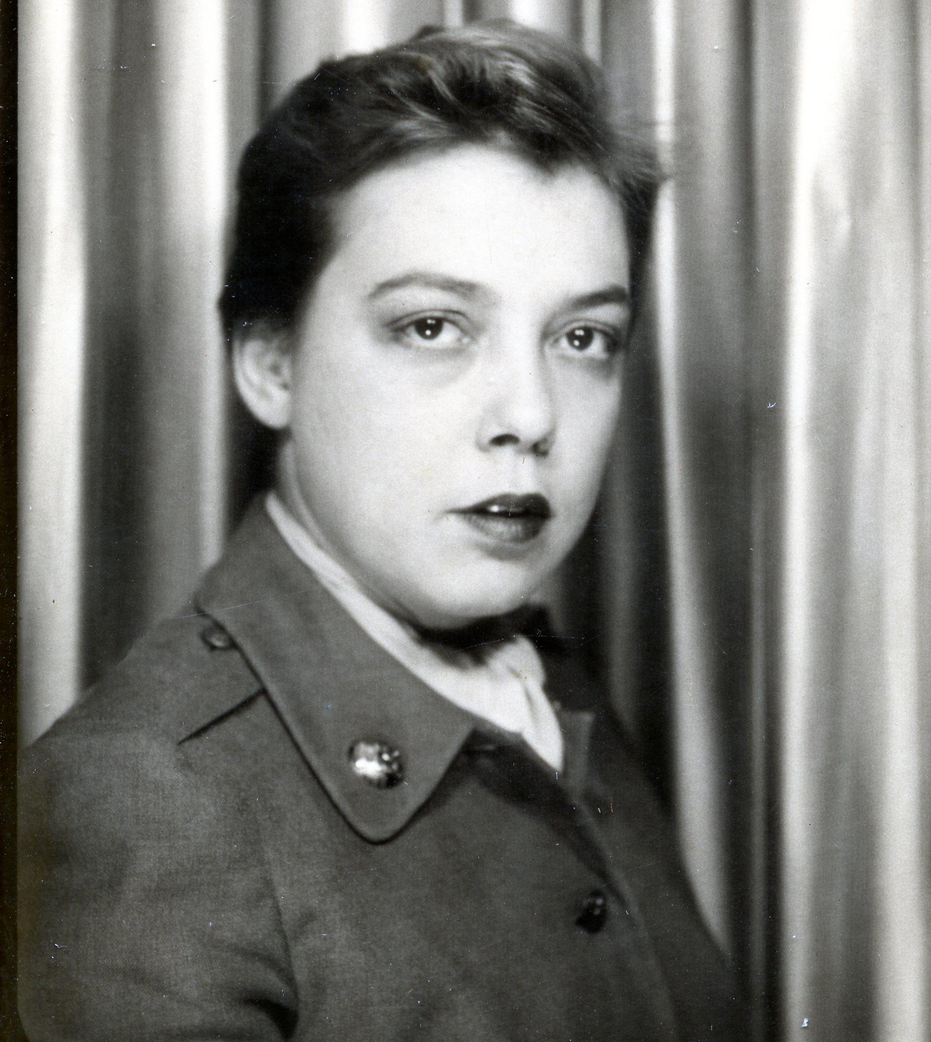 A young woman poses in a military-style uniform against a curtain backdrop, exuding confidence.