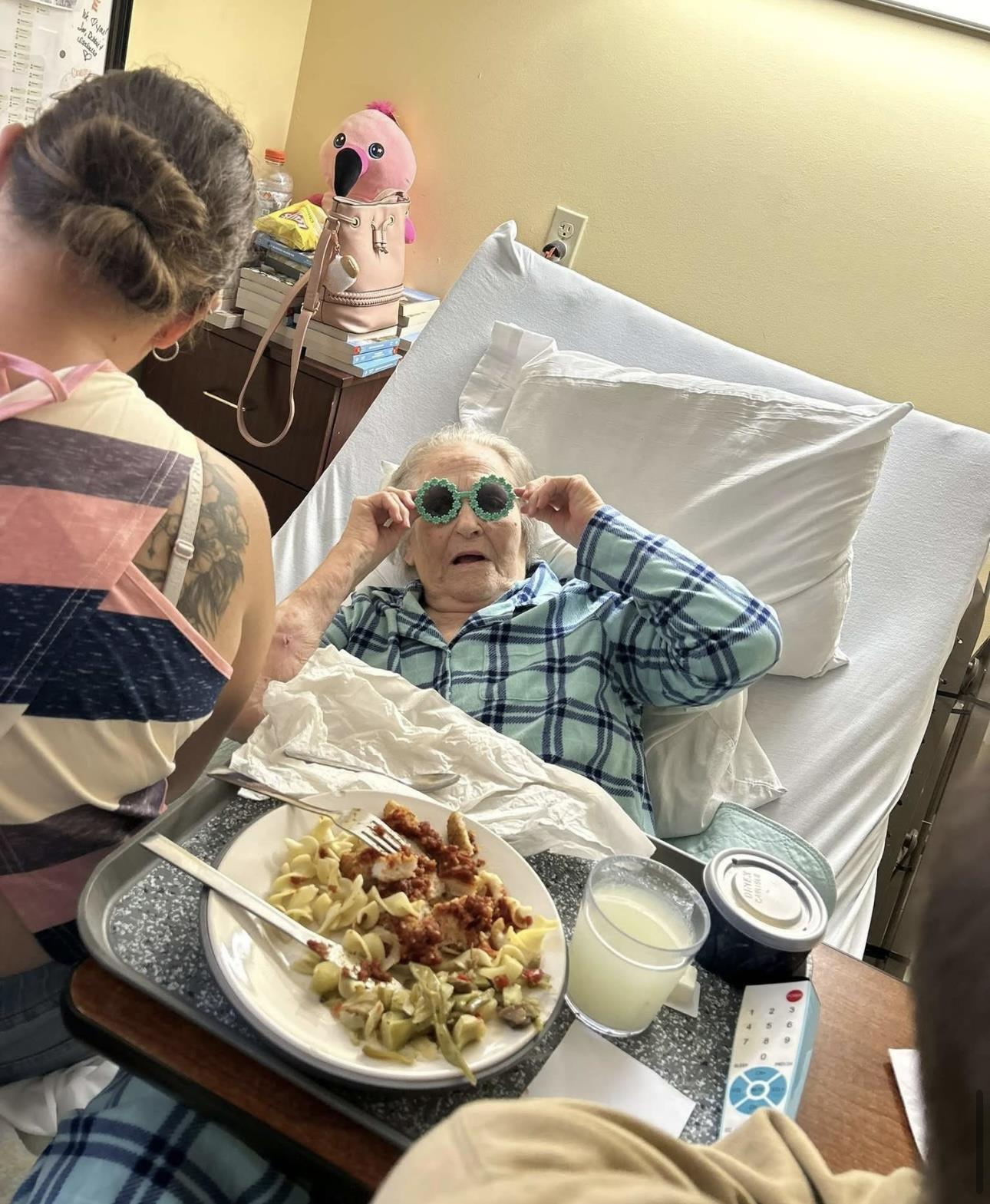 An elderly woman relaxes in bed, wearing sunglasses and enjoying a meal with others nearby.