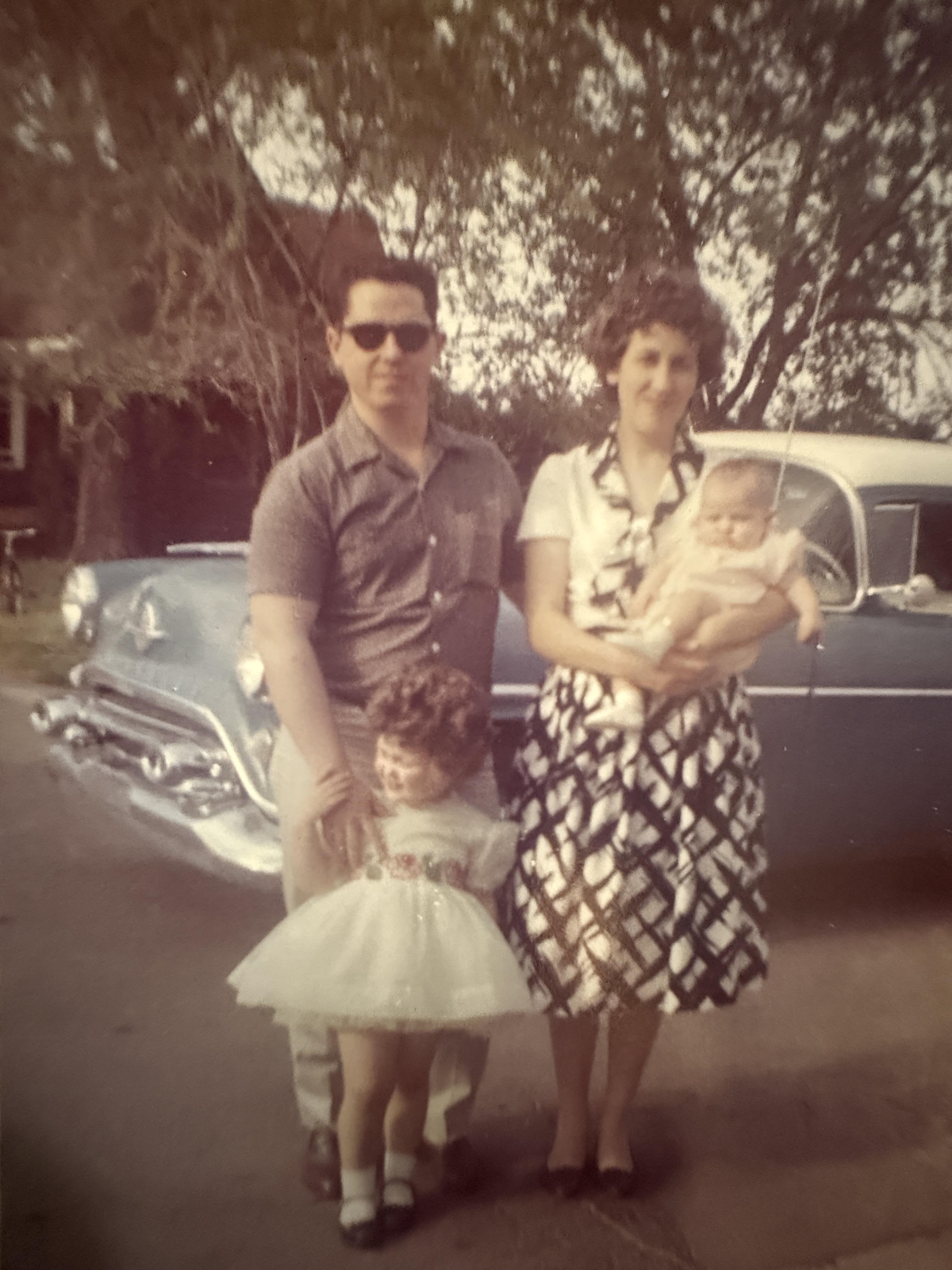 A couple stands proudly with their children in a nostalgic setting beside a classic car.