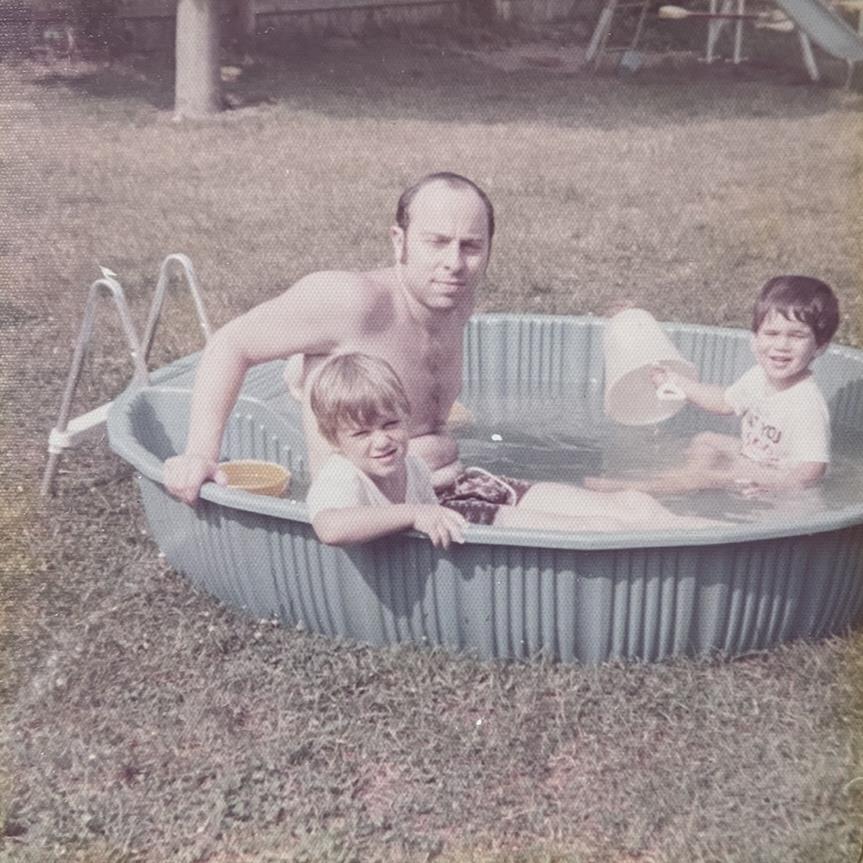 Two kids are playing in a small pool with their father on a sunny day in the backyard.