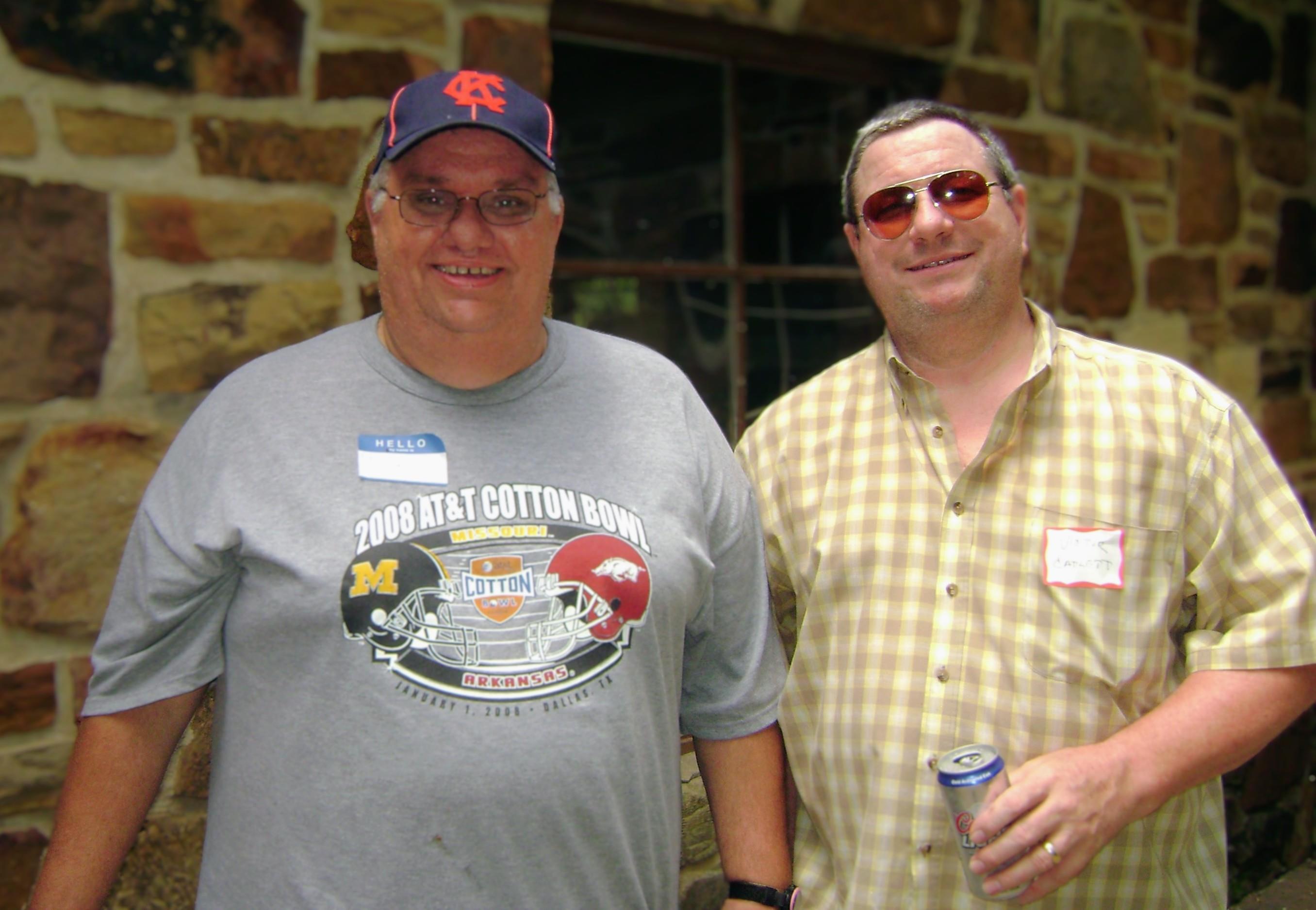 Two men stand side by side, smiling and holding drinks outdoors during a social gathering.