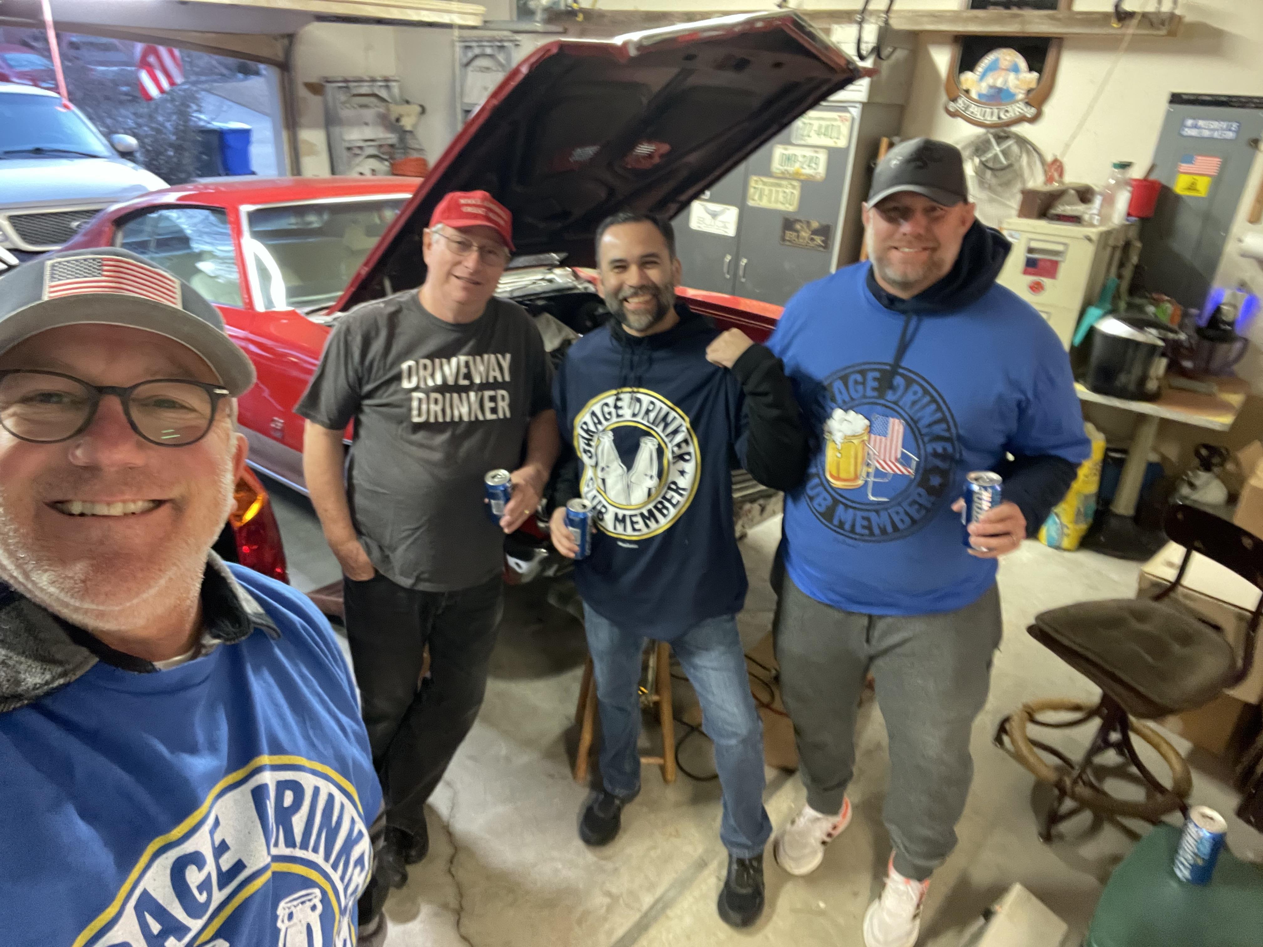 Four friends gather in a garage, holding drinks and sharing laughs while examining a classic car.