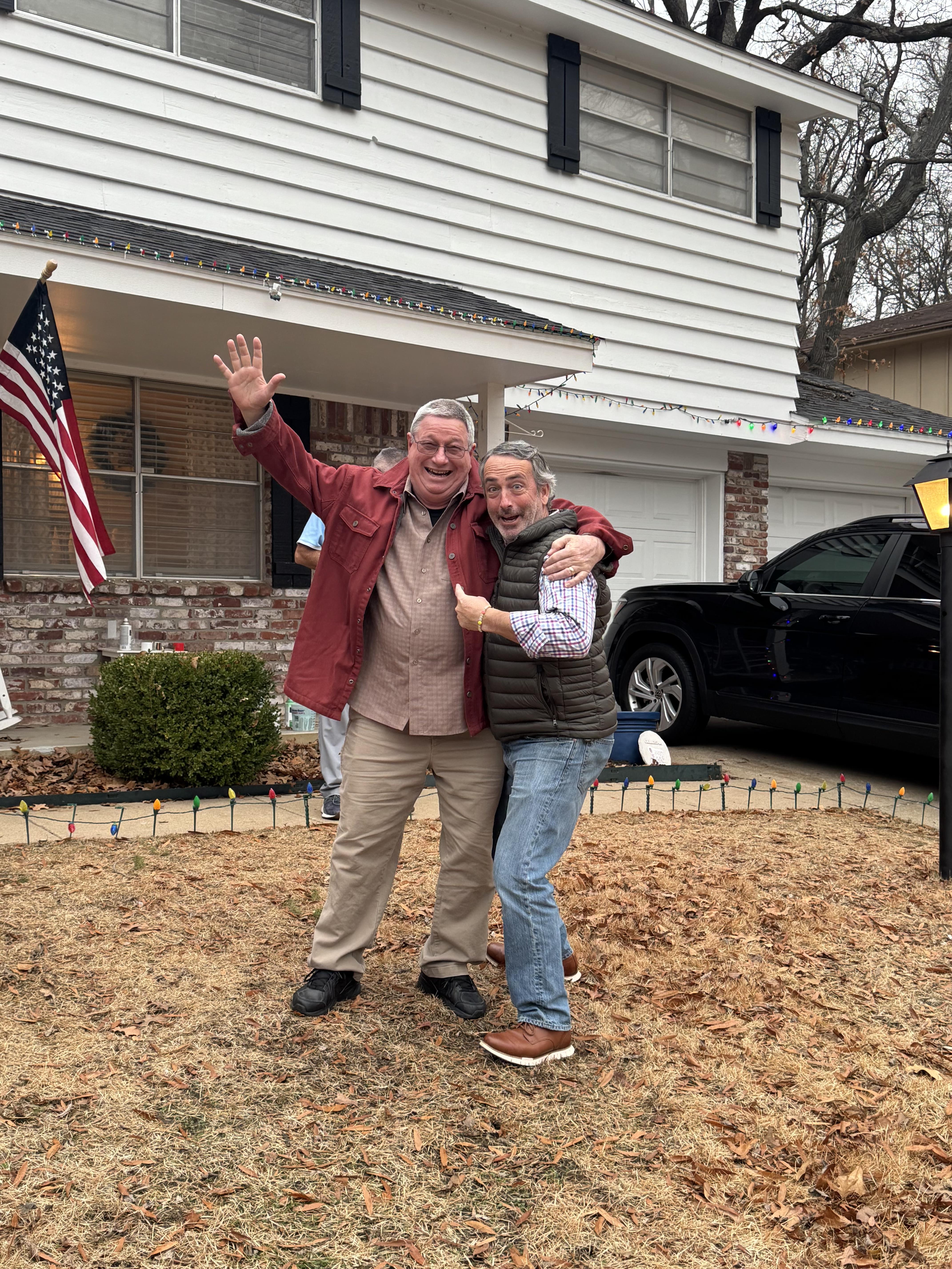 Two men embrace and smile broadly in front of a house adorned with holiday decorations.
