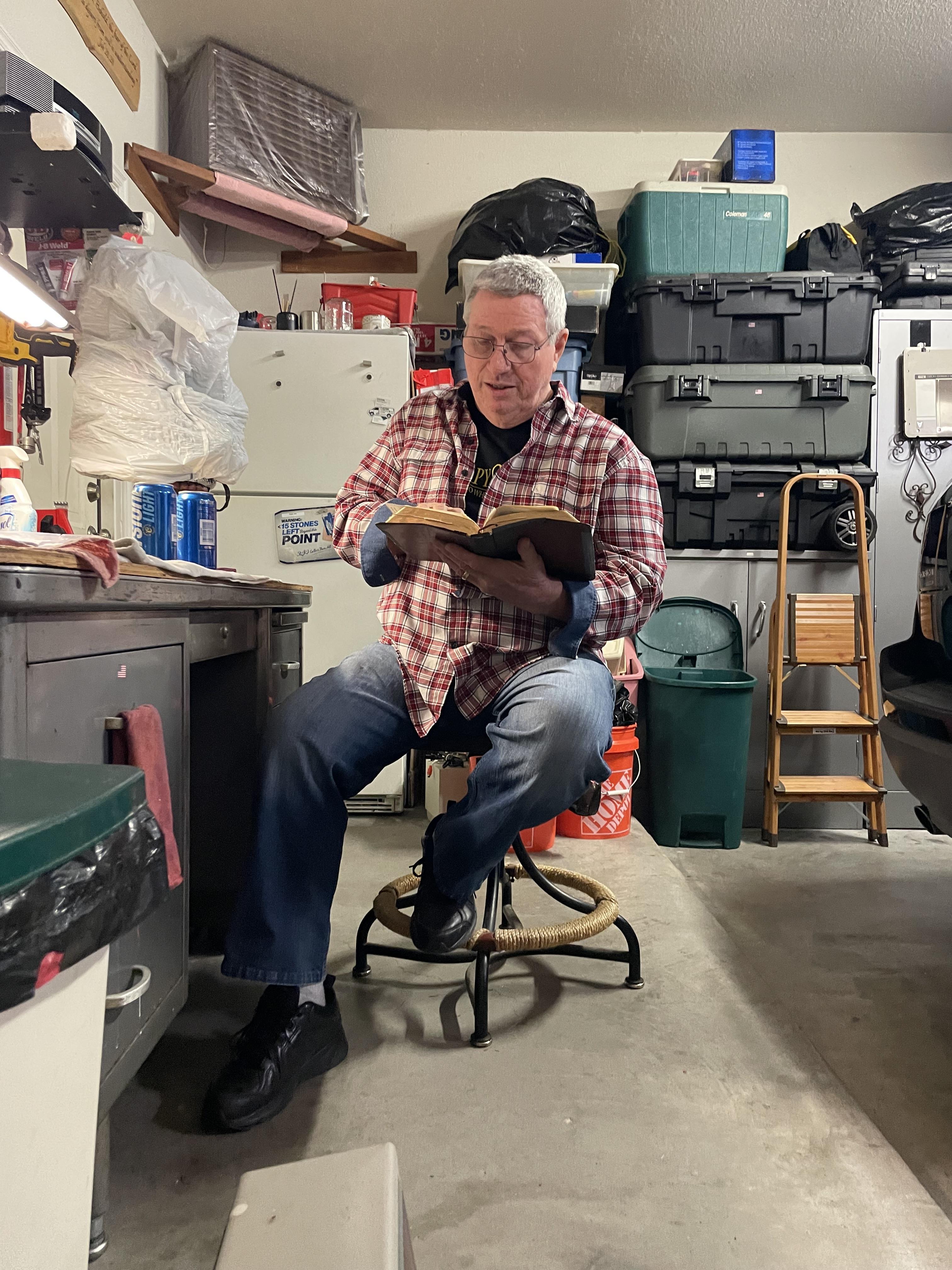 A man enjoys reading a book in a cluttered garage, surrounded by tools and various supplies.