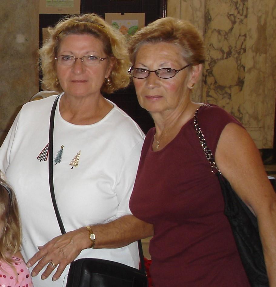 Two middle-aged women stand closely together, smiling, amidst a classic architectural interior.