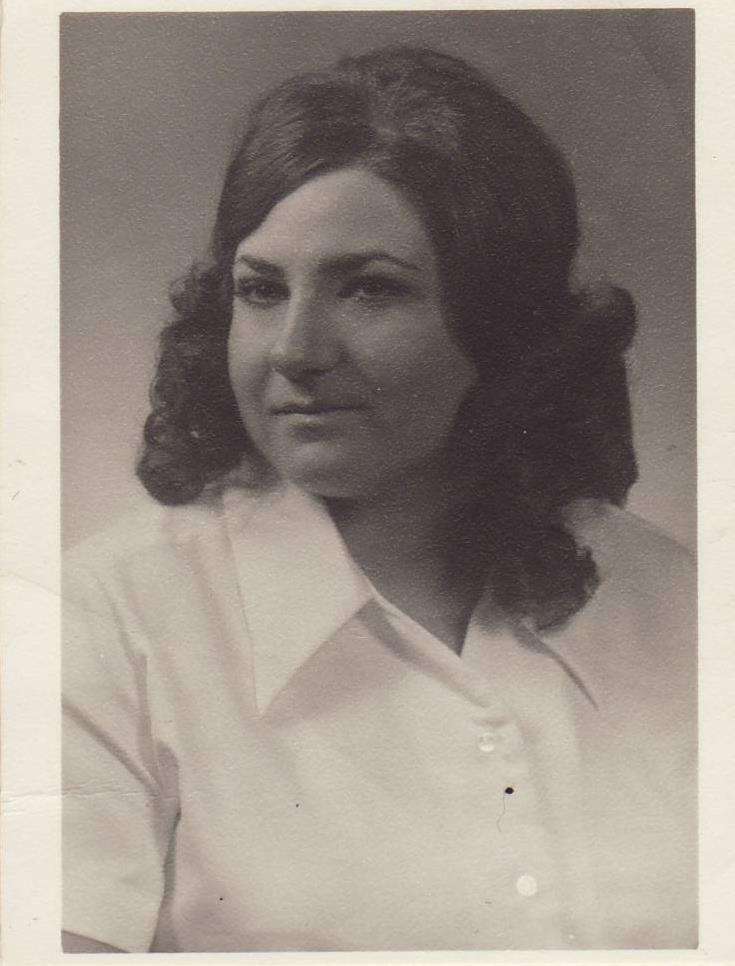 A woman with curly hair poses in a vintage studio, wearing a white blouse and smiling softly.