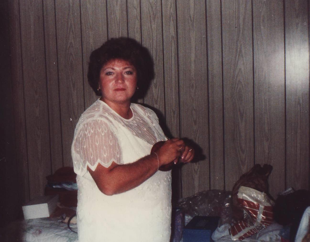 A woman poses indoors, dressed in light clothing, with a relaxed demeanor in a cozy setting.