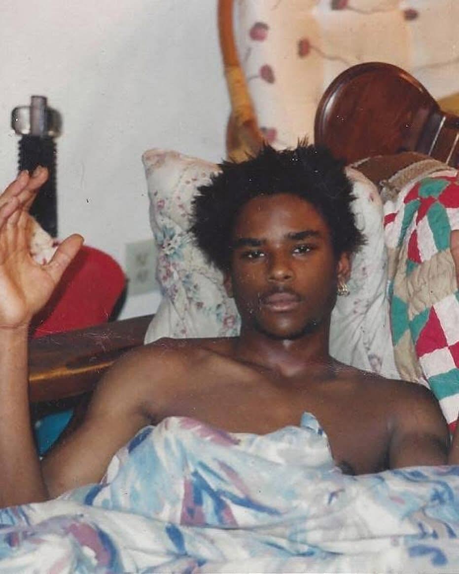 A young man with tousled hair lounging in bed, resting his hands casually in the air.
